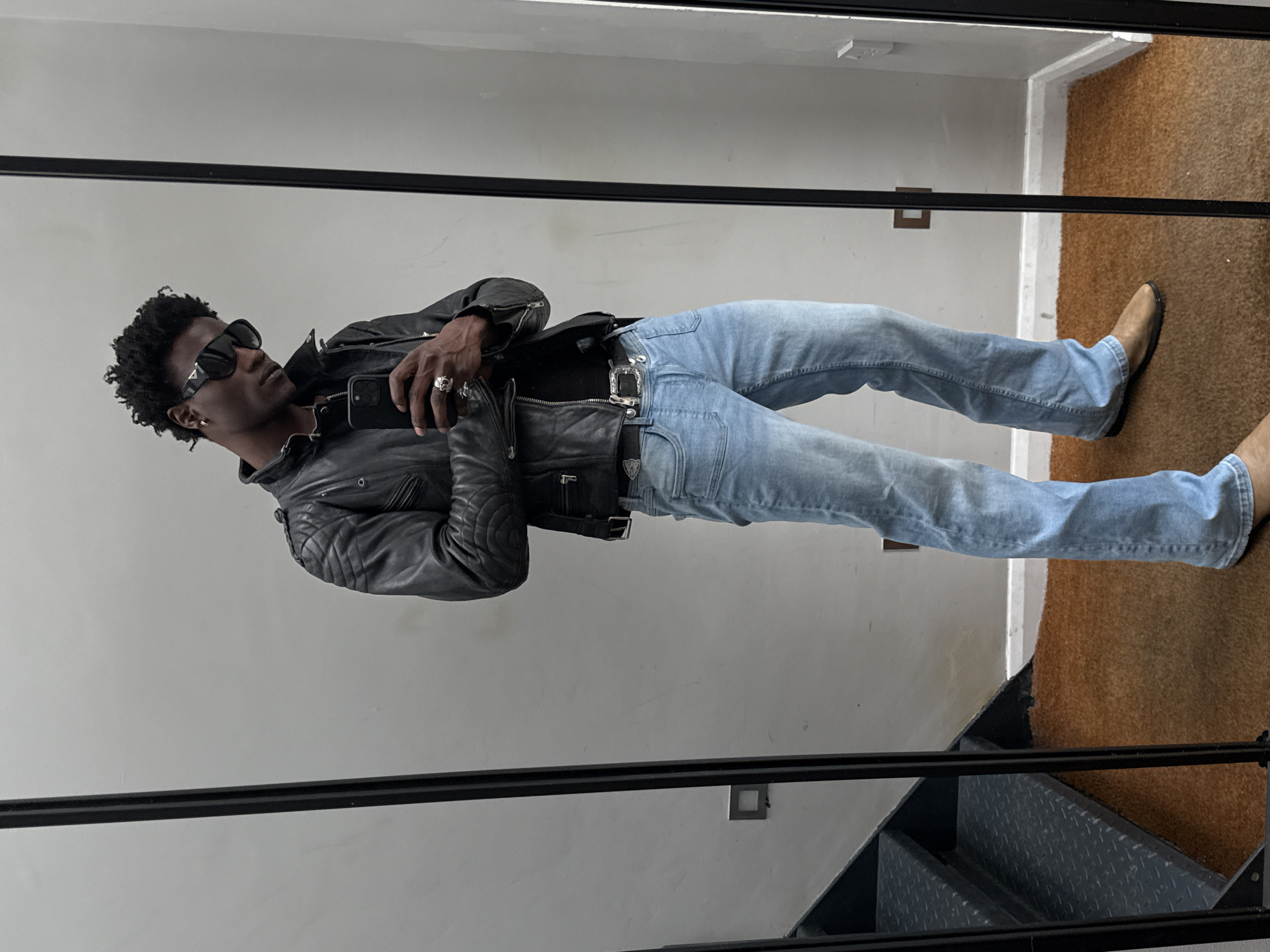 The image is a selfie of a Black man with an afro hairstyle, wearing a black leather jacket, light blue jeans, and tan-colored shoes. He is wearing sunglasses and jewelry on his fingers. He is holding a black phone and standing in what appears to be a narrow space with light-colored walls and a staircase visible in the foreground. A mirror is reflecting the man and the surroundings. The floor is covered with a brownish-orange carpet. The location is likely indoors, possibly an apartment or building interior in the city of London, United Kingdom.