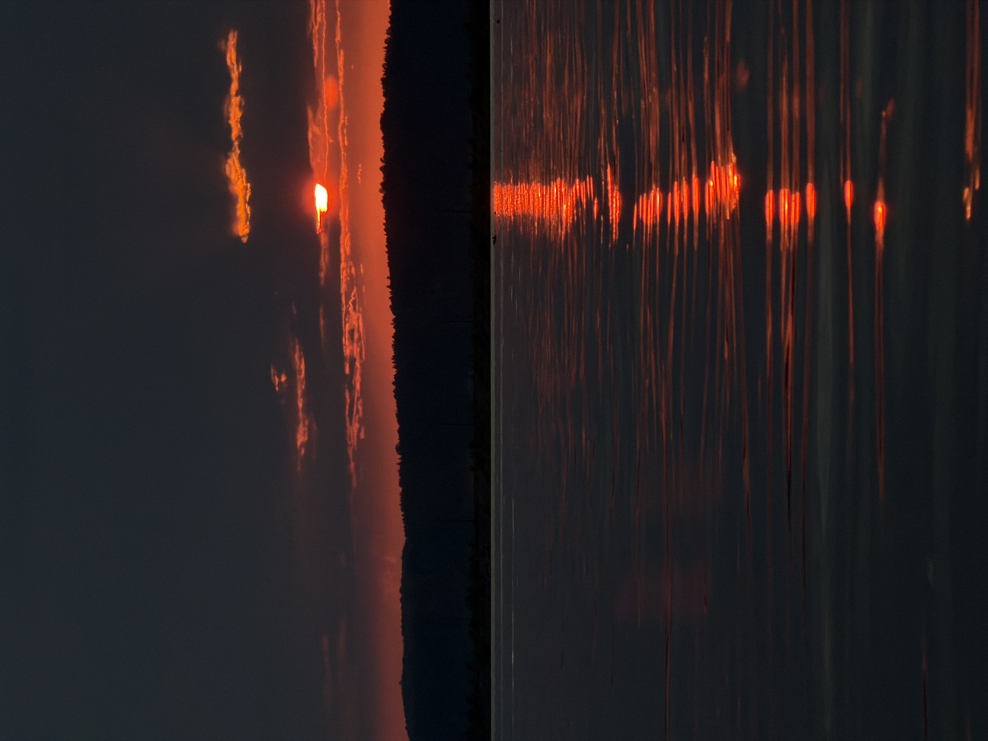 The image shows a sunset over the Kama River in Chaikovsky, Russia. The sun, a bright orange orb, is partially obscured by dark clouds. Its reflection shimmers on the water's surface, creating a trail of light. A silhouetted treeline stretches across the horizon, casting a dark shadow over the landscape. The overall scene is one of serene beauty, with the vibrant colors of the sunset contrasting with the dark, muted tones of the surrounding environment. The water is calm, with gentle ripples distorting the reflection of the sky. No people are visible in the image.