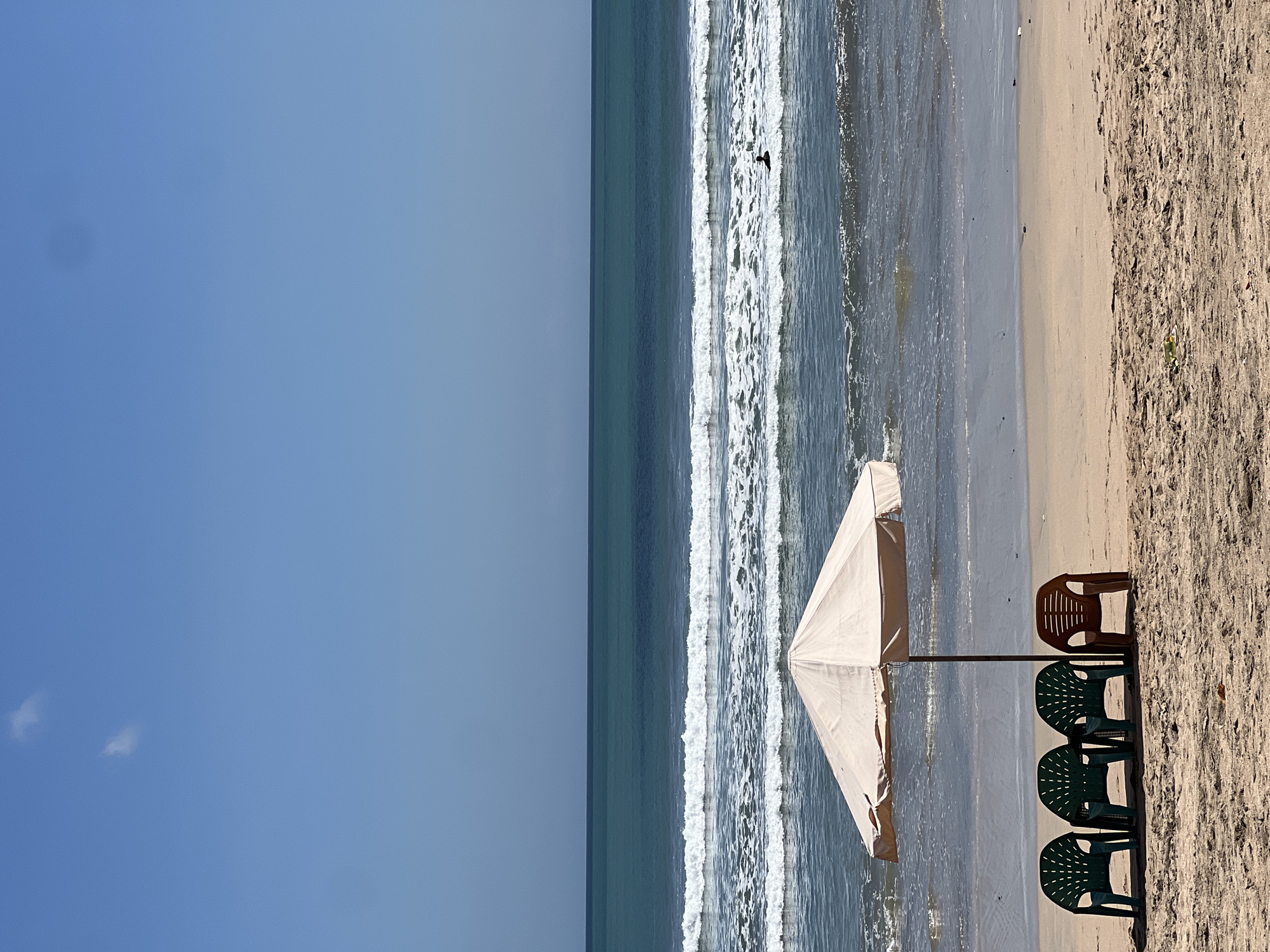 The image depicts a serene beach scene in Kuta, Indonesia, under a clear blue sky with two small clouds. In the foreground, four plastic chairs are arranged on the sandy beach. Three of the chairs are dark green, while one is a lighter brown. These chairs are positioned beneath a large, light-colored beach umbrella, providing shade. The ocean stretches out in the background, with gentle waves breaking onto the shore. A lone surfer is visible in the water, waiting for a wave. The overall environment is calm and inviting, characteristic of a tropical beach destination.
