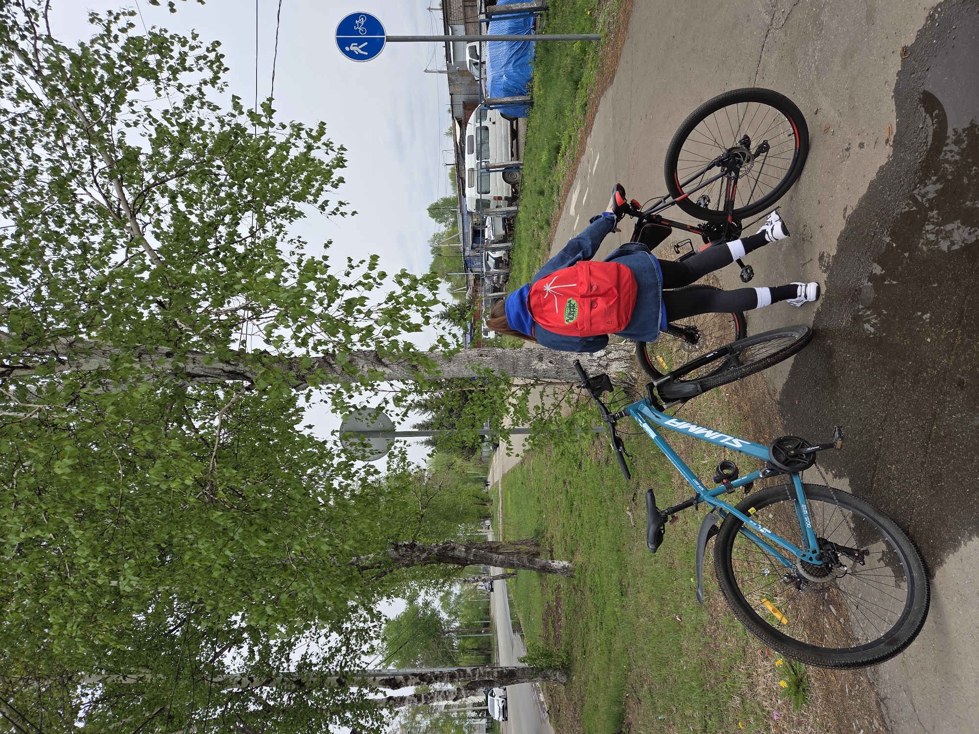 A young person wearing black leggings, white sneakers with black and white striped socks, a blue hooded sweatshirt, and a denim jacket, is seen from the back, with a red backpack. The person is standing on a paved surface near a tree, next to two bicycles. One bike is teal and the other is black with red accents. To the right of the person, there is a puddle on the pavement. In the background, there's a combination of trees, grass, a road, and buildings. A blue and white bicycle and pedestrian sign is also present. The location appears to be Birobidzhan, Russia.