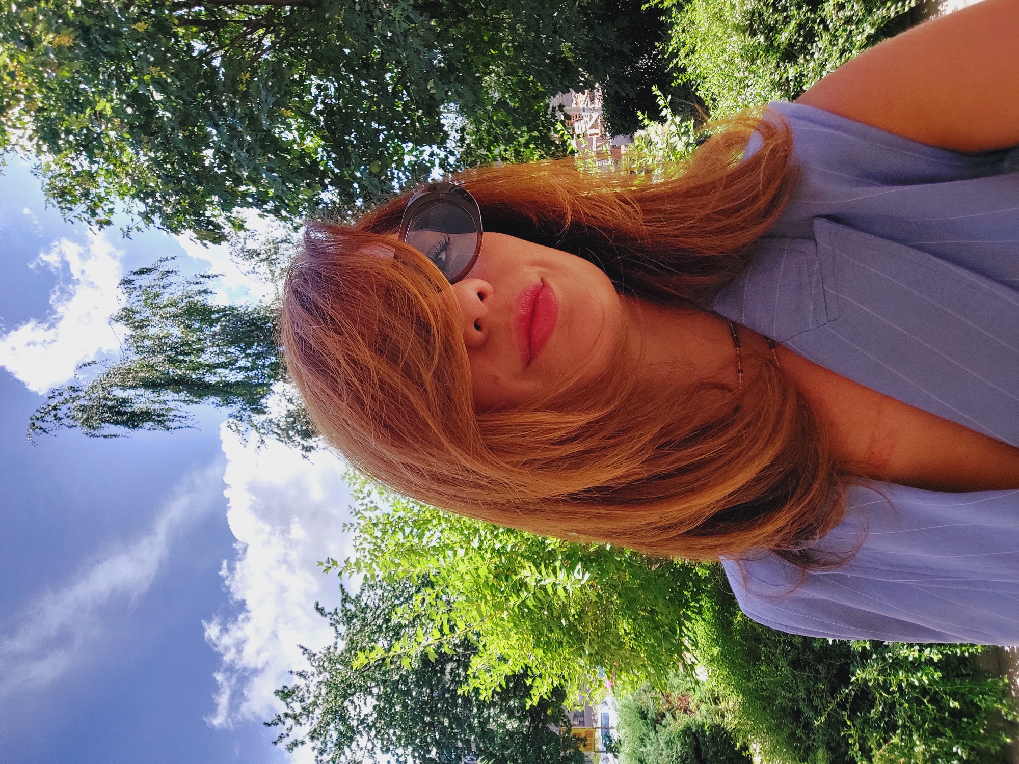 A woman with auburn hair and sunglasses takes a selfie outdoors on a sunny day. She is wearing a light blue pinstripe vest. Behind her, lush green trees and a blue sky with white clouds are visible. The background suggests a park or garden setting, possibly in the city of Sofia, Bulgaria, given the architecture and vegetation that are typical of the region.