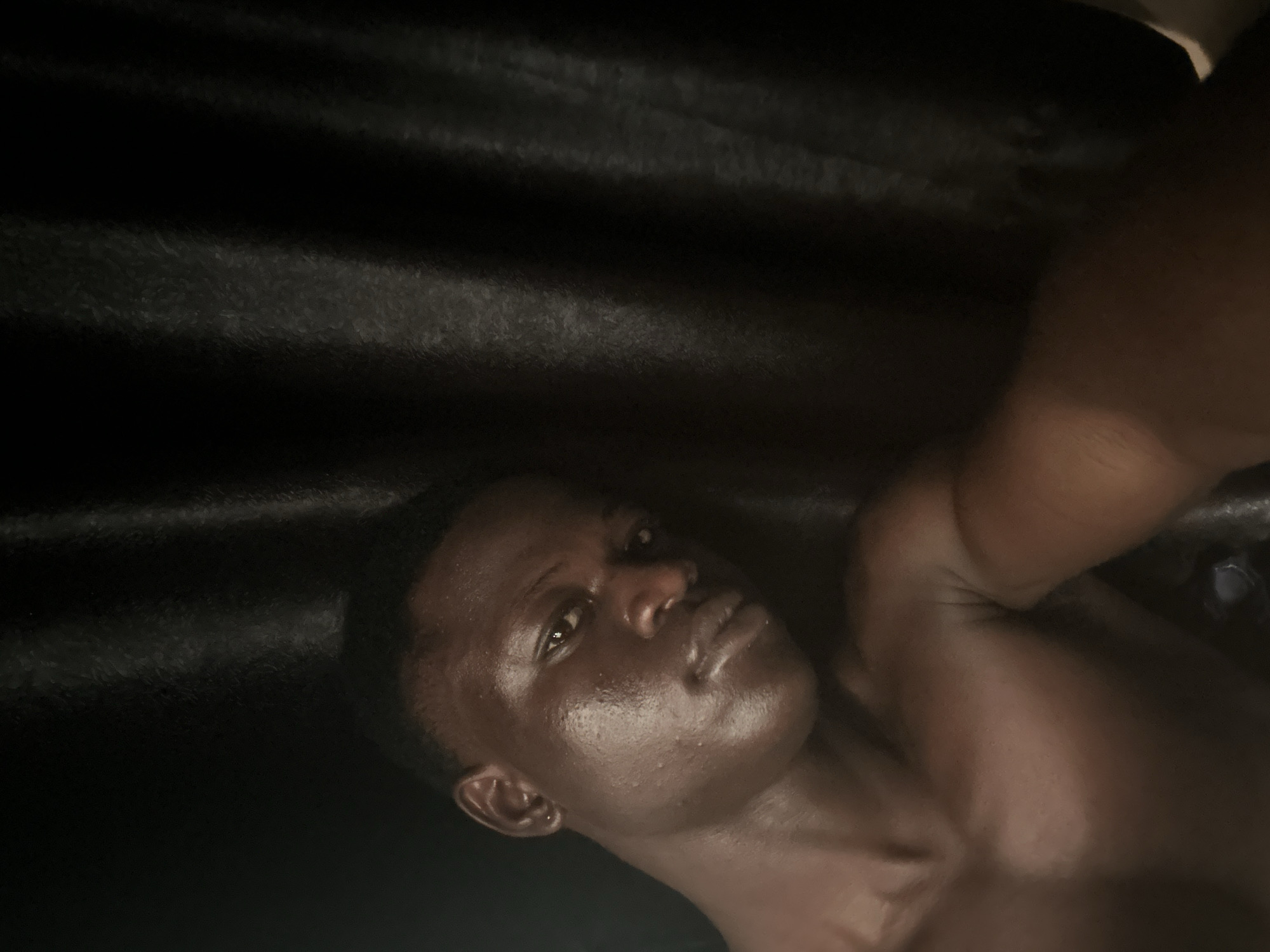 This is a close-up, low-angle, selfie-style portrait of a young Black man. He is shirtless and looking towards the camera, with his head tilted slightly to his left. His skin appears to be shiny, possibly from sweat or oil. The background is a dark, textured material, likely a curtain or drapery, with vertical folds. The lighting is dim, casting shadows and highlighting the contours of his face and upper body. The man has short hair and appears to be in a relaxed pose. There are no other people or objects clearly visible in the frame besides the man and the background material. The image does not contain any visual information that would suggest its location in Badagry, Nigeria, or any other specific geographical place.