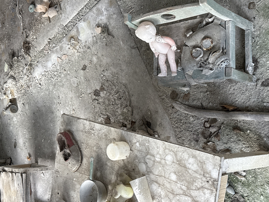 The image depicts an abandoned interior, likely a classroom or playroom, within the city of Pripyat, Ukraine. The scene is dominated by concrete surfaces covered in significant dust and debris, indicating a prolonged period of disuse and decay.
In the left foreground, a light-colored, rectangular table is present, its surface similarly caked with dust and marked with circular stains. On this table, from left to right, are: a small, light-colored metal saucepan with a pale green handle; a yellowed, irregularly shaped object; a white, cylindrical object; and a weathered, reddish-brown and grey child's shoe, appearing worn and dusty.
To the right of the table, on the floor, is an accumulation of detritus including fragments of plaster or concrete, small stones, and organic matter. A long, slender piece of weathered wood lies diagonally on the floor, extending towards the lower left.
In the lower right portion of the image, a light green wooden child's chair is visible. Resting on the seat of this chair is a pale-skinned doll, facing away from the viewer, with its head tilted to the right and its limbs positioned as if reclining. Its clothing appears simple and possibly lace-trimmed at the neck. Partially obscured beneath the doll, also on the chair, is a decomposed or damaged gas mask, showing rusted metal components and disintegrating material.
The overall environment suggests abandonment, with no human figures present. The objects, primarily children's items, are left in situ amidst the general decay.