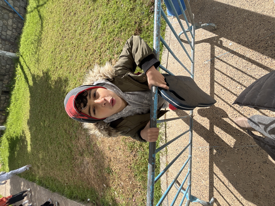 The scene depicts an outdoor daytime environment in Meknes, Morocco, viewed from a high-angle perspective looking downwards. A young male, appearing to be a boy or adolescent, is the primary subject, positioned behind a weathered blue metal railing. He has dark, curly hair and wears a gray and red patterned beanie, a gray hooded sweatshirt, and an olive-green jacket with a fur-lined hood. He grips the top bar of the railing with both hands and looks directly towards the camera with his mouth slightly open in a puckered expression. In his left hand, he holds a dark-colored flip-flop or sandal hanging over the railing. A red shoe is visible on his right foot.
The blue metal railing features a horizontal top bar and multiple vertical and diagonal supports. Behind the railing, the ground transitions from a light-colored paved surface with dark aggregate to a strip of dry, light-brown earth, followed by a vibrant green grassy lawn. In the far background, beyond the lawn, a stone or concrete wall is visible.
To the left in the background, partial figures of at least two other individuals are discernible, one wearing white pants and light shoes, and another with visible legs in dark pants and white shoes, both moving away from the camera. The foreground shows the lower portion of the photographer's legs and feet, clad in light-colored boots and dark pants, standing on a light-colored paved surface similar to the one behind the railing. The scene is brightly illuminated by direct sunlight, casting sharp, elongated shadows of the railing and figures onto the ground.