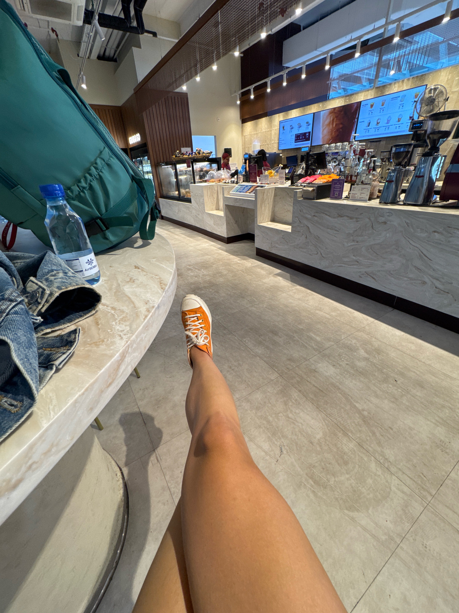 The image shows a first-person perspective of a person sitting at a marble-topped table in what appears to be a modern coffee shop, possibly in Palma de Mallorca, Spain. The person is wearing orange Converse sneakers and has their legs crossed, with one leg visible from the knee down. A partially open green backpack and a bottle of water are on the table to the left. In the background, there is a coffee bar with electronic menus displayed on screens, coffee machines, and various pastries. The floor is tiled.