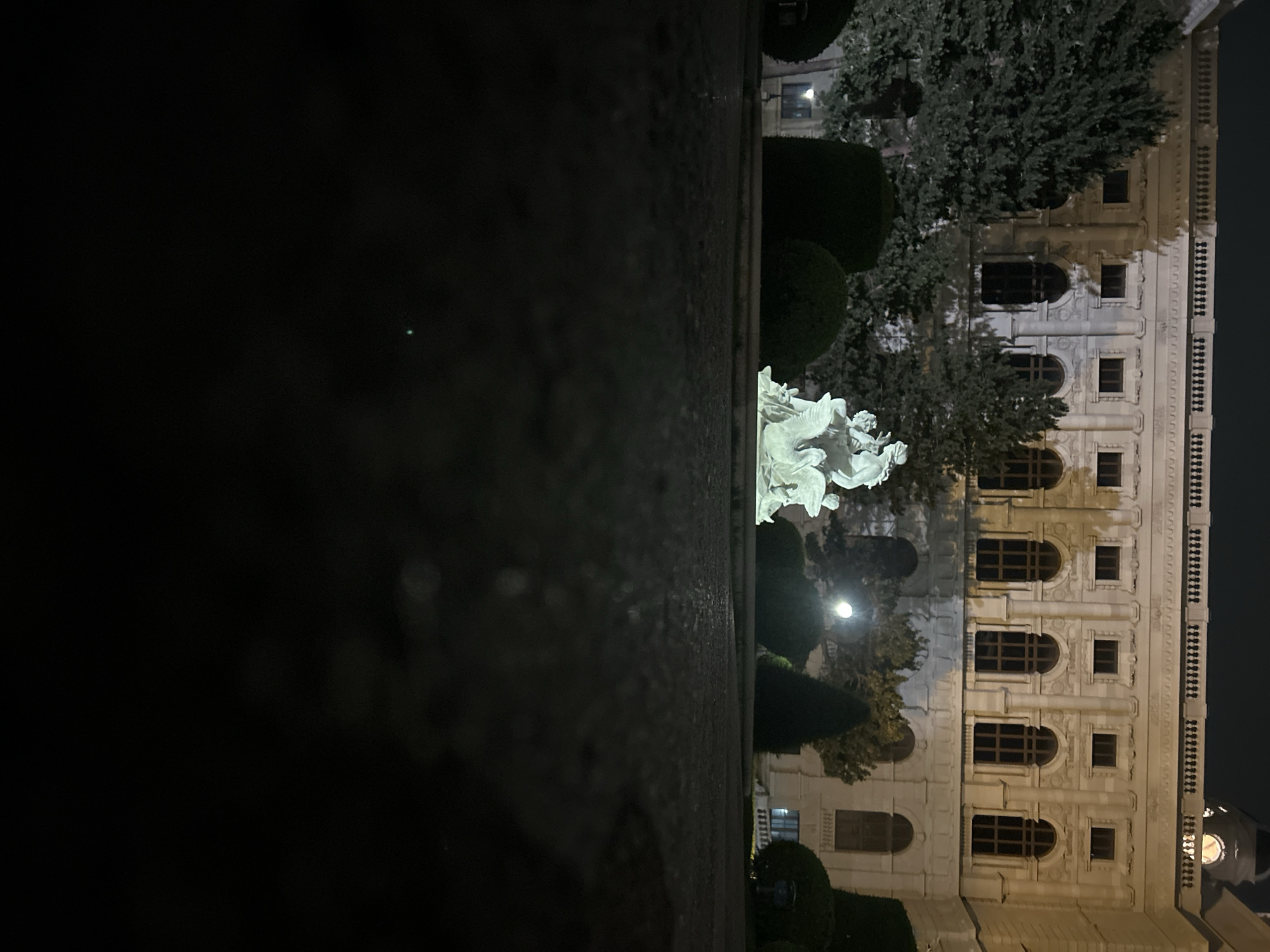 The image shows a night scene in front of the Naturhistorisches Museum (Natural History Museum) in Vienna, Austria. In the foreground, a dark, wet pavement is visible, suggesting recent rain or a fountain. In the middle ground, a bright white statue of a winged creature carrying figures is illuminated. Behind the statue, manicured dark green bushes and trees form a backdrop. The museum building, with its ornate facade, large arched windows, and classical architectural details, is prominent in the background. The building is lit up, highlighting its grandeur. The sky is dark, indicating nighttime.