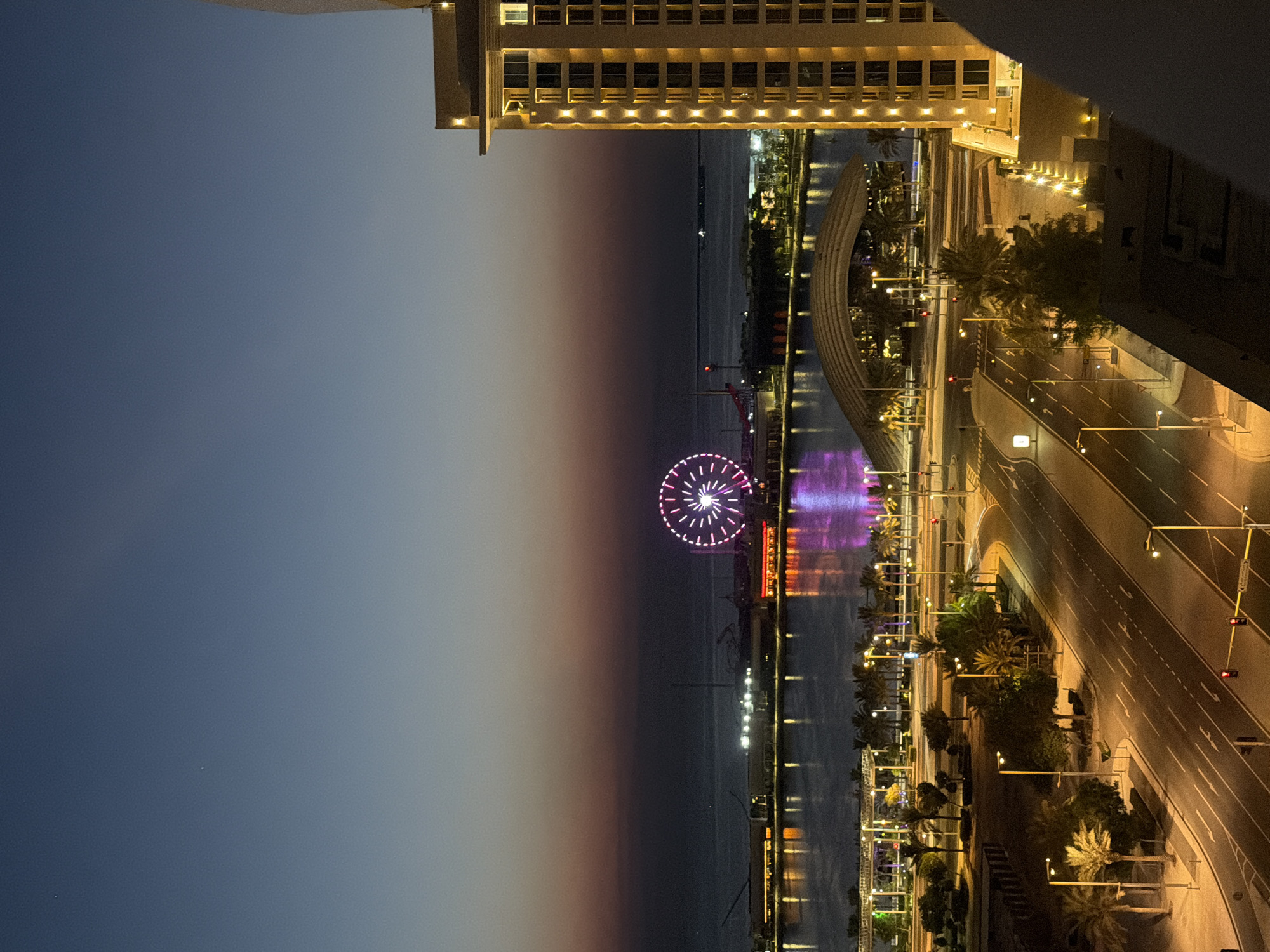 An aerial, eye-level, long shot captures a twilight view of a waterfront city. The sky transitions from deep blue at the top to a dusky purple and orange near the horizon, where the sea meets the land. A large Ferris wheel, illuminated with pink and white lights, stands prominently in the distance over the water. Along the waterfront, buildings are lit up, and their reflections shimmer on the calm water. A modern highway with multiple lanes curves through the foreground, lined with streetlights and palm trees. A tall, lit hotel building dominates the right side of the frame. The scene is devoid of people and appears to be a cityscape at dusk.