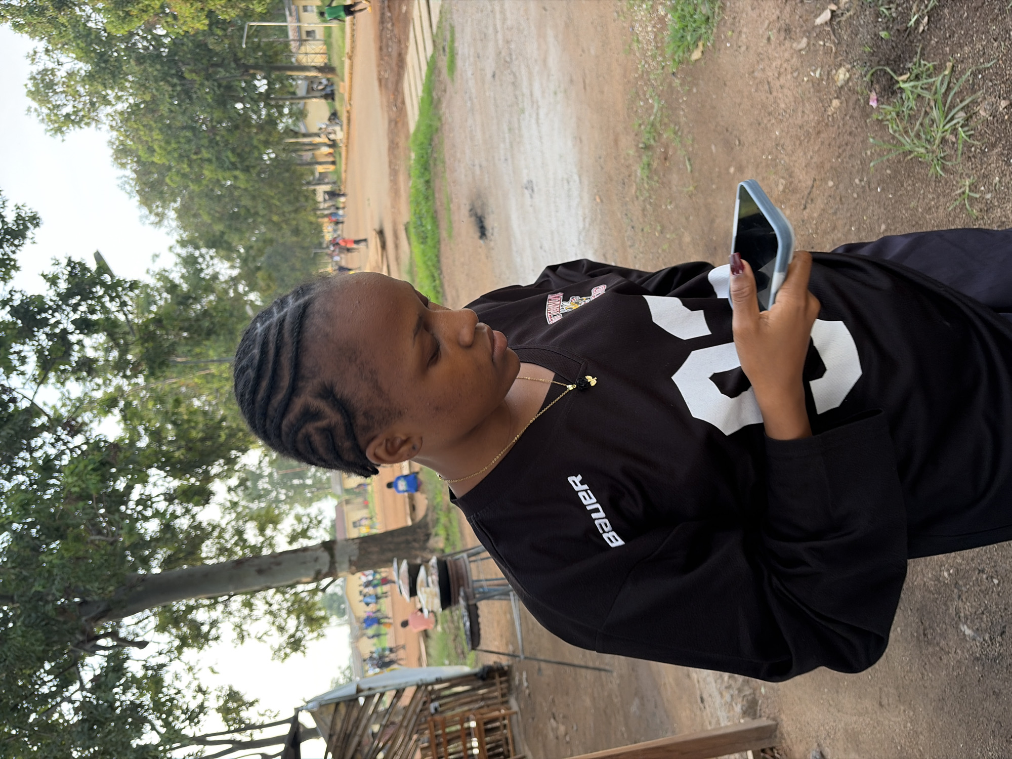 An outdoor, medium shot captures a young woman in Rafin Par, Nigeria, as she looks down at her smartphone. The woman, with her hair in intricate braids, wears a black jersey with white lettering and a small gold necklace. She is holding a light blue smartphone in her right hand. The background shows a dusty street with scattered trees and buildings, and several people in the distance. A wooden structure is visible on the left side of the image. The lighting suggests it is late afternoon or early evening.