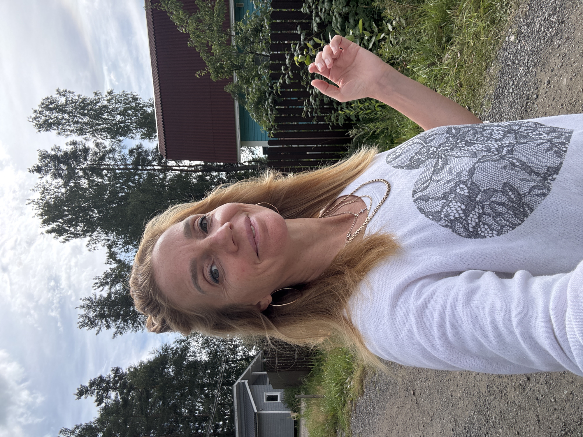 A medium shot captures a blonde Caucasian woman in her 40s standing outdoors on a gravel path. She is wearing a white long-sleeved shirt with a large gray lace heart graphic on the chest. She has on two gold necklaces and large hoop earrings. Her hair is pulled back in a messy bun with blonde strands framing her face. She is smiling at the camera with her right hand raised slightly, fingers extended. In the background, there are two houses, one a light blue house with a red roof, and a smaller gray house with a darker roof. A wooden fence separates the properties. Tall evergreen trees and deciduous trees line the background, with a cloudy sky above. The overall setting appears to be a rural or suburban area in Steklyannyi, Russia, indicated by the architectural style of the houses and the surrounding vegetation.