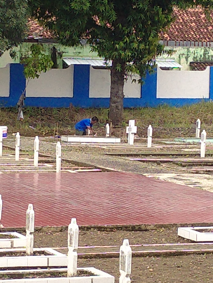 The image captures a daytime scene within a cemetery or burial ground in Karanganyar, Indonesia. In the mid-ground, a lone individual, dressed in a blue short-sleeved shirt and dark pants, kneels beside a grave marker. The person appears to be meticulously cleaning or tending to the grave with a small object in their hands.
The cemetery is characterized by numerous white grave markers. Some are simple, upright rectangular posts, while others are distinctly cross-shaped, indicating a Christian burial ground. A prominent red-tiled pathway or area cuts across the mid-ground, separating sections of graves. The ground between the graves consists of earth and sparse, dry vegetation.
In the background, a large, leafy green tree stands tall, its foliage obscuring part of a blue and white painted wall or fence. Beyond this wall, faint glimpses of buildings with orange-tiled roofs and light green walls are visible through the trees, suggesting a surrounding residential or village area. A white bucket or container is visible to the far left in the foreground.
The lighting is soft and diffused, indicating an overcast day or perhaps early morning/late afternoon, without harsh shadows. The overall atmosphere suggests quiet devotion and upkeep. There is no discernible text visible on any of the grave markers or other objects in the image.