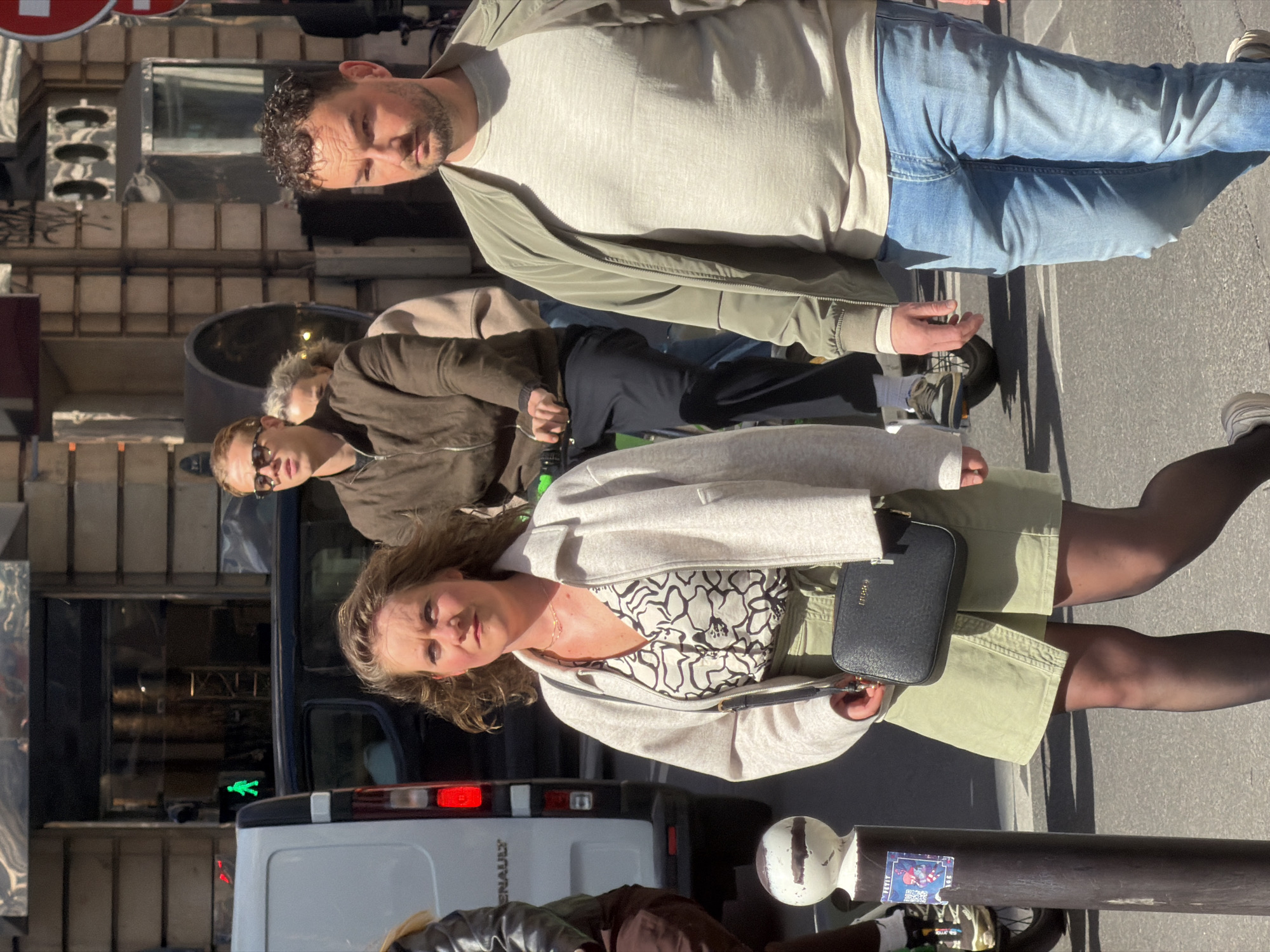 The image shows a street scene in Paris, France. There are four individuals: a man in a grey shirt and blue jeans standing at the top of the image; a man wearing sunglasses and a brown jacket; a man wearing a tan jacket; and a woman in a white sweater, patterned shirt, and green skirt in the foreground. The woman is carrying a black purse. All of the people are standing on the sidewalk. A white Renault van is partially visible in the background, along with a pedestrian traffic signal. The environment includes Parisian architecture, a street light pole, and pedestrian traffic.