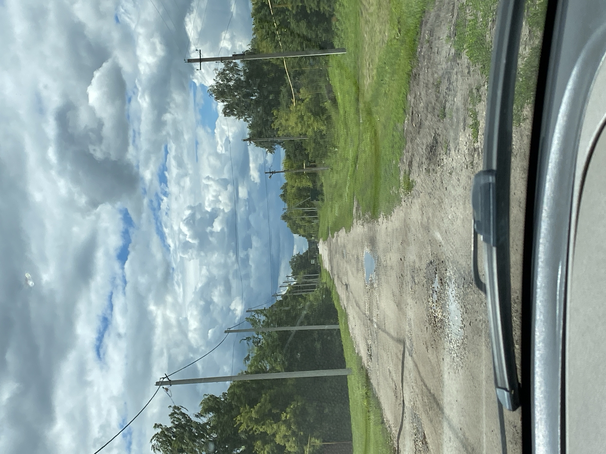 An eye-level, wide-angle shot looks down a rural, unpaved road in Nadezhdino, Russia. The road is rutted and has several puddles, suggesting recent rain. Telephone poles with wires line both sides of the road, interspersed with trees and green grass. The sky is cloudy with patches of blue sky visible. The foreground shows the windshield and wiper of a vehicle.