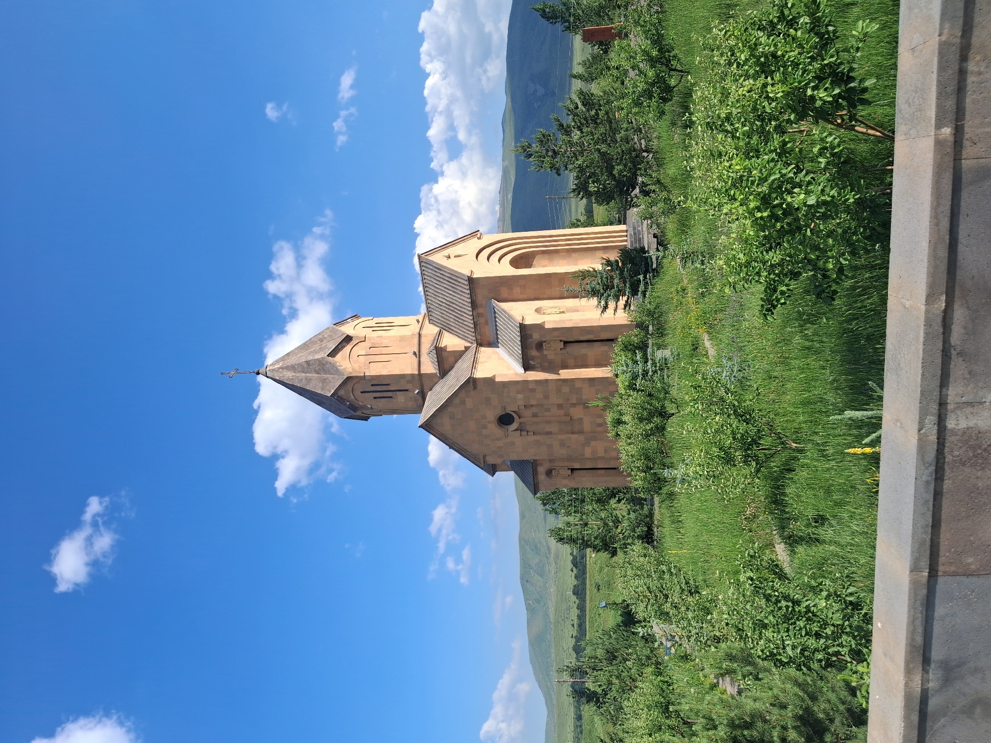 A beige-colored church with a conical roof topped by a cross stands prominently in a green landscape. The church features arched windows and doorways, with carved details. Surrounding the church is a well-maintained area with lush green grass, small trees, and bushes. In the background, rolling hills are visible under a bright blue sky dotted with white clouds. A stone wall is in the foreground of the image. The church is located in Jermuk, Armenia.