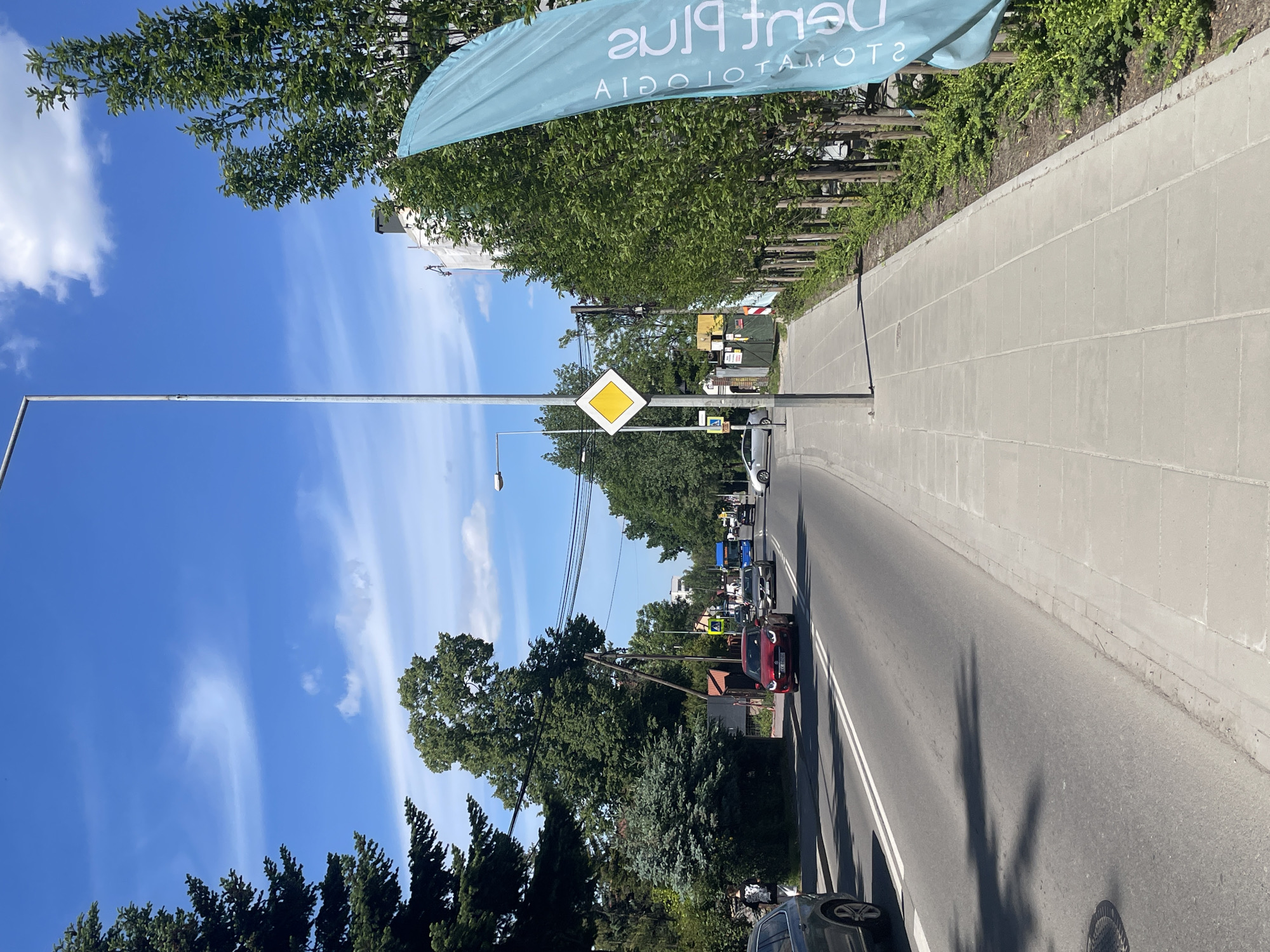 The image shows a street in an urban environment. A yellow diamond-shaped sign is visible on a pole near the road. Several cars are traveling along the street, and the sidewalk is visible on the right side. A blue banner with the words "Dent Plus Stomatologia" is partially visible in the upper portion of the frame. The sky is blue with scattered clouds. The buildings are surrounded by green trees and shrubbery.