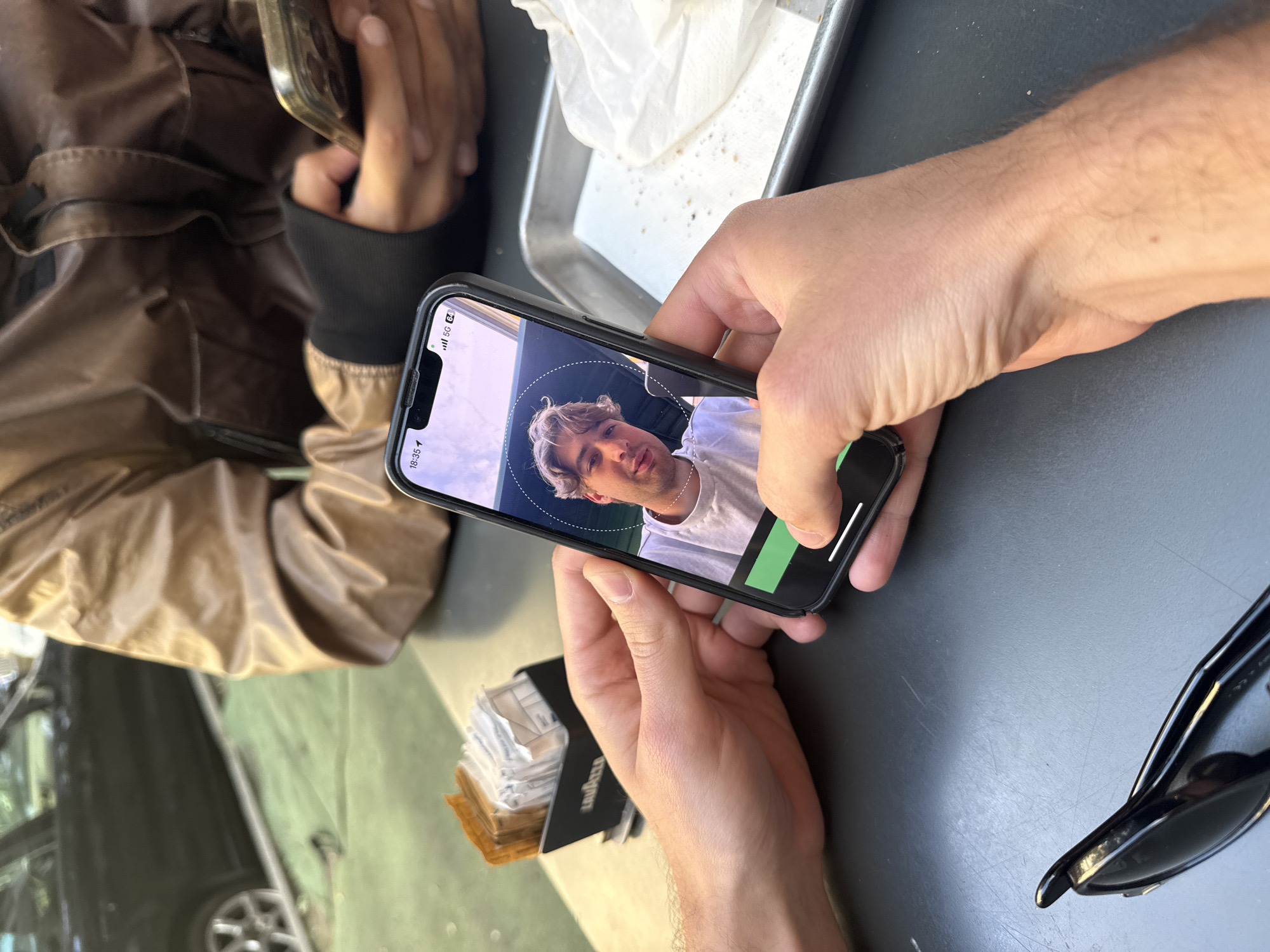 Here is a forensic description of the image:
A smartphone displays a man's face within a white circular frame, potentially indicating facial recognition technology is in use. The phone is held by fair-skinned hands with visible veins. A stack of Lavazza sugar packets sits to the left of the phone. A pair of black sunglasses rests on a gray surface to the right. In the background, parts of a brown jacket and another smartphone are visible. A metal tray holds a white napkin with crumbs. Through a window, a partial view of a vehicle with a multi-spoke wheel can be seen. While the location in the city of Milan, Italy cannot be confirmed, the presence of Lavazza sugar packets could suggest an Italian setting.