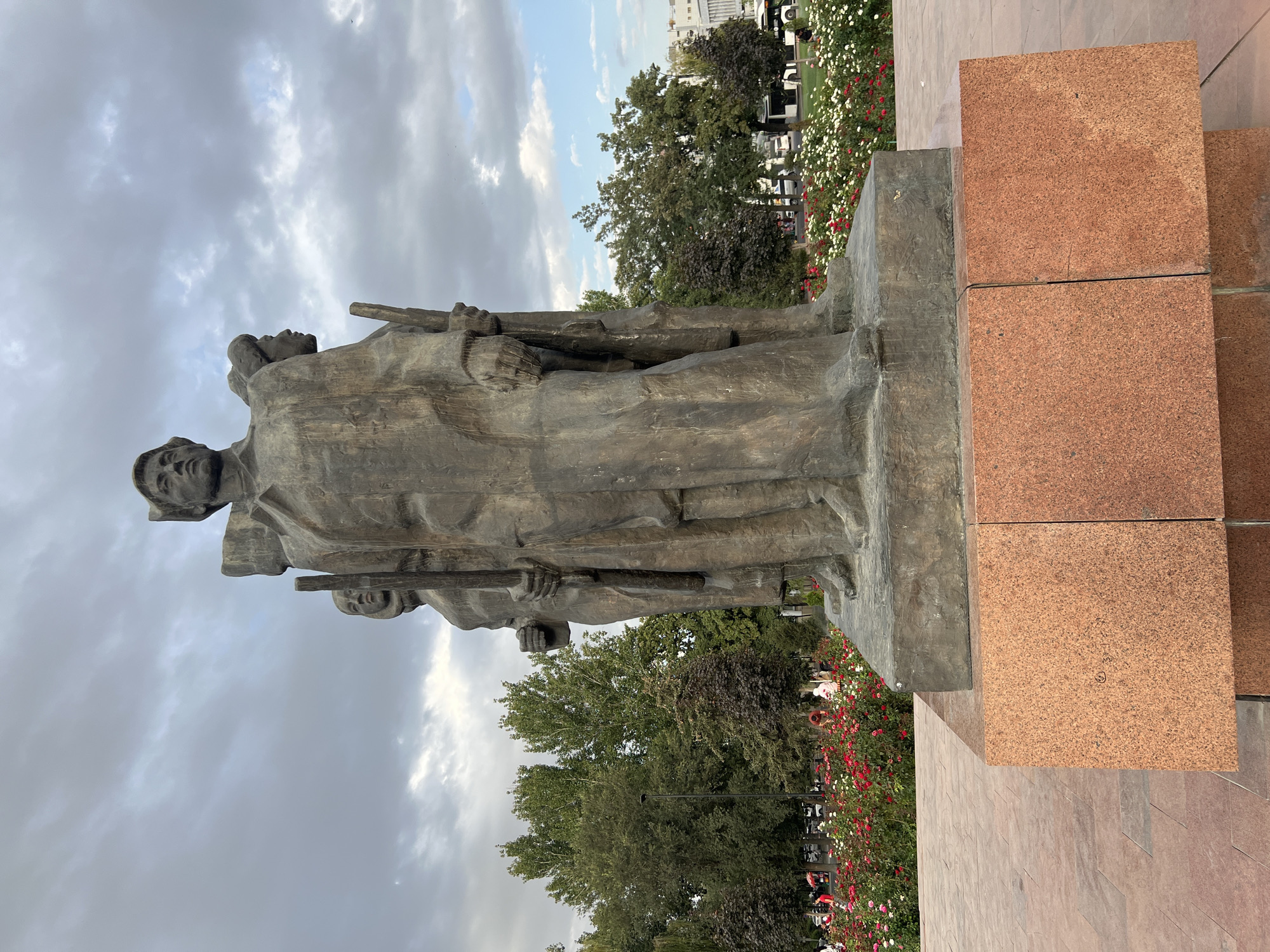 The image shows a bronze sculpture of three figures, two men and one woman, standing together. The central figure is a man in a long coat, holding a rifle. To his left and right are two other figures, also holding rifles. The sculpture is mounted on a rectangular granite pedestal. In the background, there are trees, a flower bed with red and white roses, and a cloudy sky. A street with cars and people can be seen beyond the flower bed. The sculpture is located in Ala-Too Square, Bishkek, Kyrgyzstan.