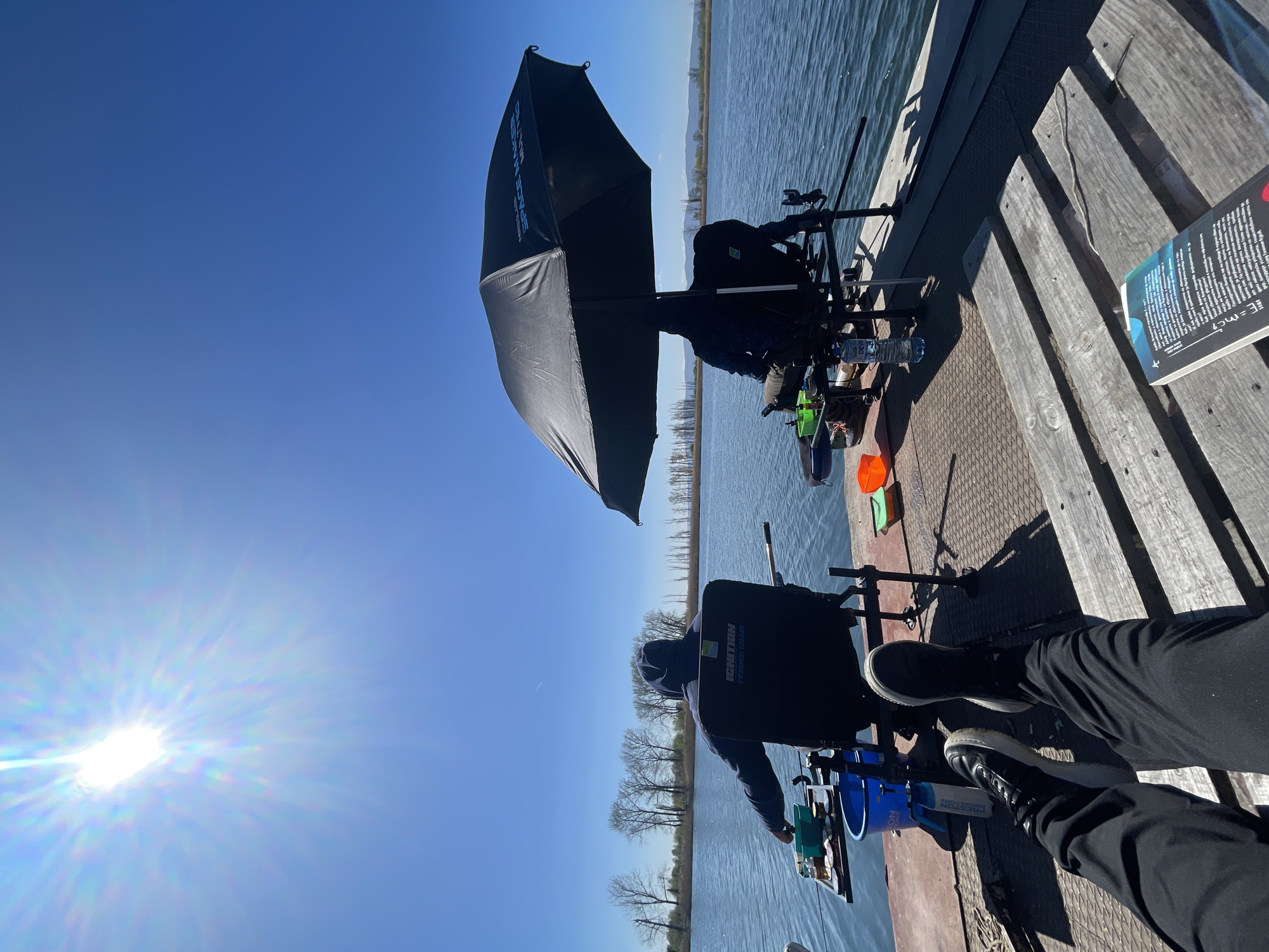 The image depicts two individuals fishing on a wooden platform near a body of water, likely a lake or river. One person is seated under a large black umbrella. The second person is shown with their feet resting near a book on a wooden surface. The sky is clear blue, and the sun is visible. There are trees visible in the background, across the body of water. The individuals are using fishing rods and tackle, and buckets and other gear are visible. The location is most likely near a body of water in Sofia, Bulgaria.