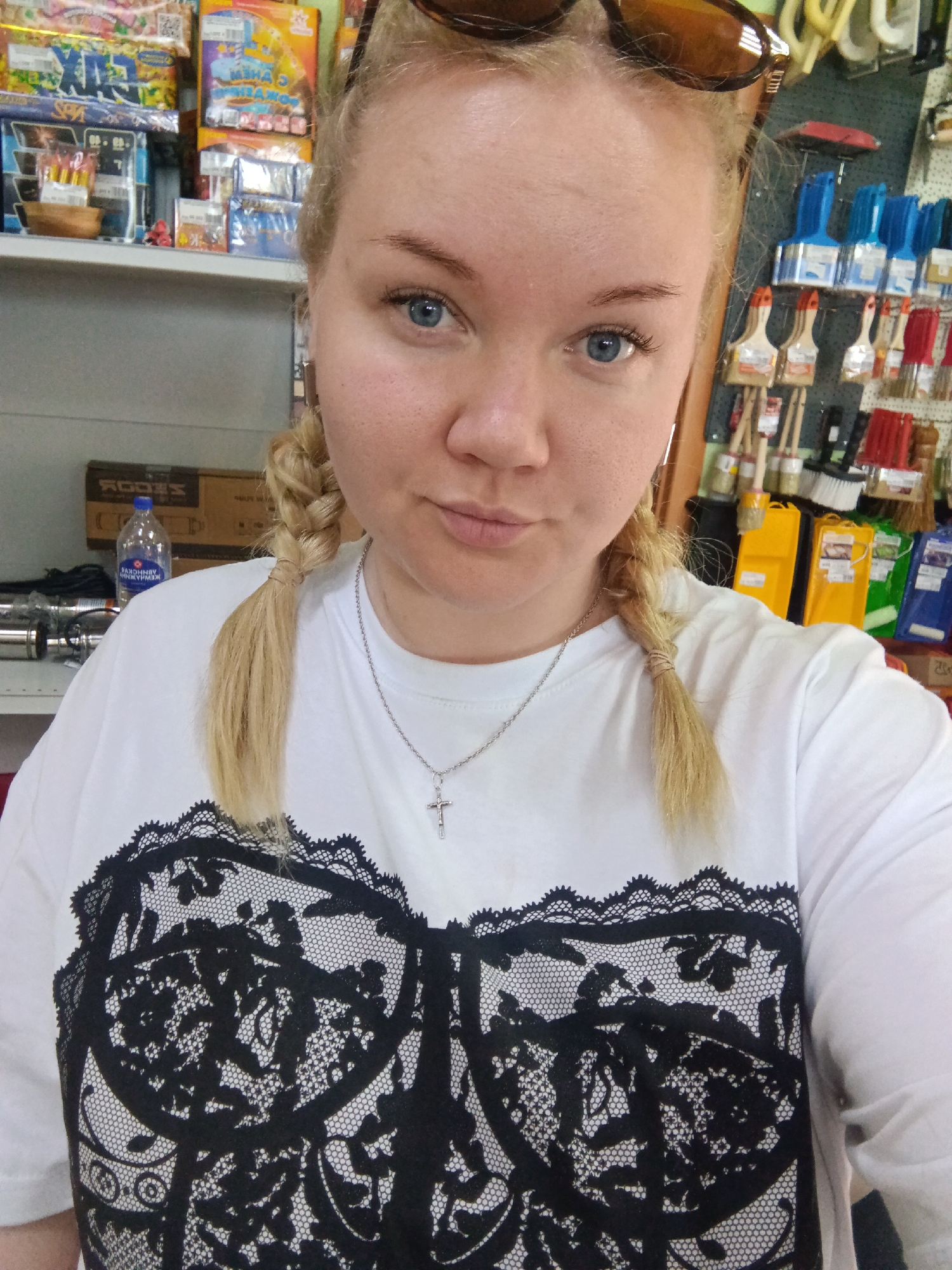 A medium eye-level shot shows a young woman with fair skin and blond hair styled in two braids. She is wearing sunglasses on her head and a white T-shirt with a black lace design resembling a bra. A silver necklace with a cross pendant is visible.
The background includes shelves stocked with various items, including paint brushes and boxes. A water bottle is also visible on a shelf.
Given that the location mentioned is Фоки, Russia, the context suggests this could be a local store selling hardware and household goods.