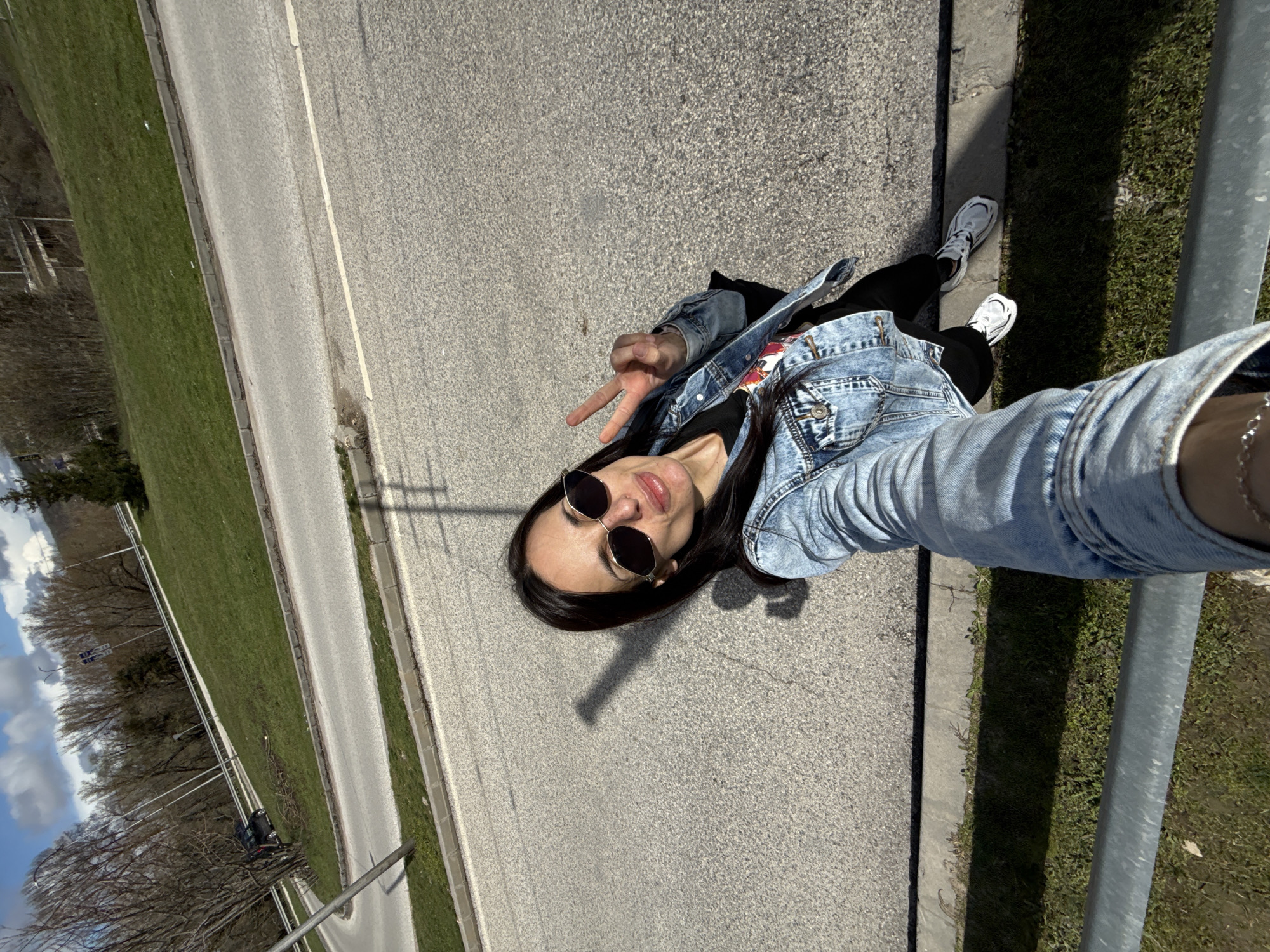 A female subject is depicted in a high-angle outdoor selfie on a clear, sunny day in Pleven, Bulgaria, specifically on **bul. "Evropa"**. The subject, of apparent young to middle age with long dark hair, is wearing hexagonal sunglasses, a light blue denim jacket over a black graphic t-shirt, black leggings or pants, and white sneakers with black accents. Her left hand is raised, displaying a two-finger "peace" gesture. Her right arm is extended, presumably holding the device capturing the image. She has slightly puckered lips.
The immediate foreground features a grey metal guardrail. Below the guardrail is a strip of green grass. The subject stands on the edge of a grey asphalt road, marked by a concrete curb and a white solid line. Shadows of the subject and a distant pole are cast prominently on the asphalt, indicating bright sunlight.
Beyond the initial road section, an elevated grassy median or embankment separates it from another parallel road segment. The background consists of a significant ascending grassy slope covered with numerous deciduous trees, mostly bare of leaves, suggesting late winter or early spring. A single evergreen conifer is visible to the right. In the far distance, faint outlines of a multi-level road structure or bridge and utility poles with road signs are discernible against a partly cloudy sky with patches of blue. A dark-colored vehicle is partially visible on an upper road section, obscured by trees on the left.