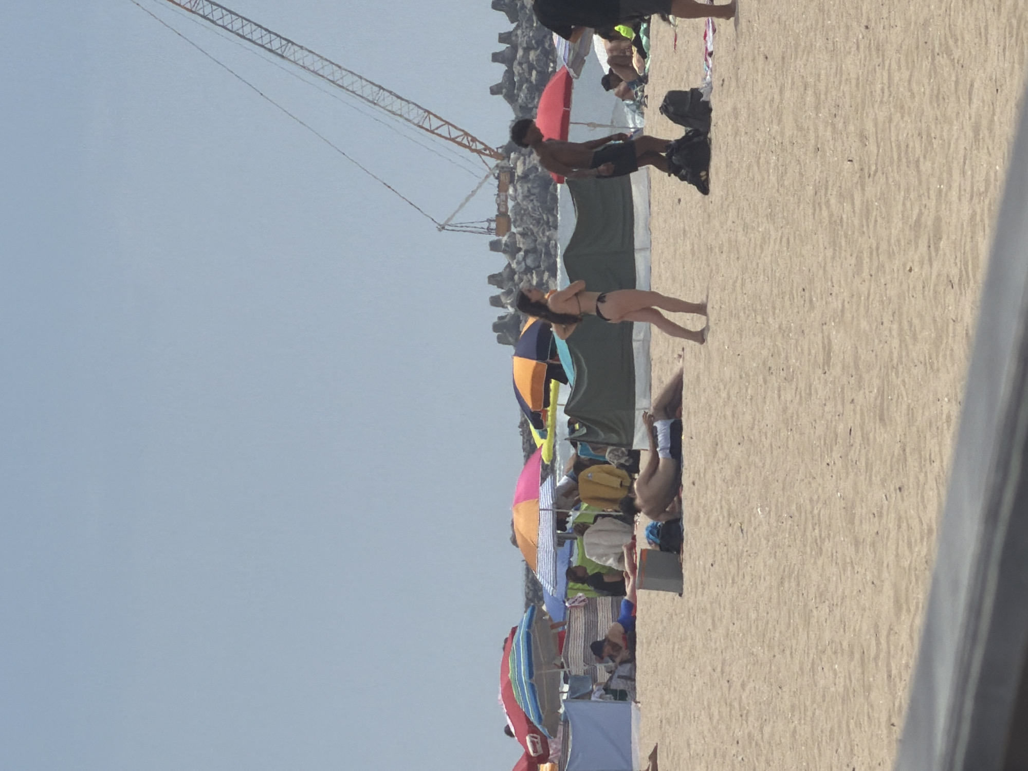 The image depicts a crowded beach scene in Espinho, Portugal, under a clear, pale blue sky. In the foreground, the sandy beach is occupied by several individuals and beach equipment. To the left, a person is lying on their stomach on a towel, wearing a blue and red bathing suit. To the right, another person is lying on their back, facing left, wearing dark swim trunks.
Mid-ground, a group of beach umbrellas are set up. A prominent orange and pink umbrella is positioned behind the person lying on their back. Further left, a red and blue striped umbrella is visible. Several tents or windbreaks are also present, providing shade and privacy for beachgoers.
In the background, a large construction crane with an orange boom is visible against the sky. The crane is situated behind a line of large, grey, concrete wave-breaking structures that extend into the ocean. People are scattered throughout the scene, some relaxing under umbrellas, others walking or standing. A woman in a black bikini is standing in the middle ground, facing right, and a man in dark swim trunks is standing to her right, facing left, with his hands on his hips. Bags and towels are visible on the sand near them. The overall impression is one of a typical busy summer day at the beach.