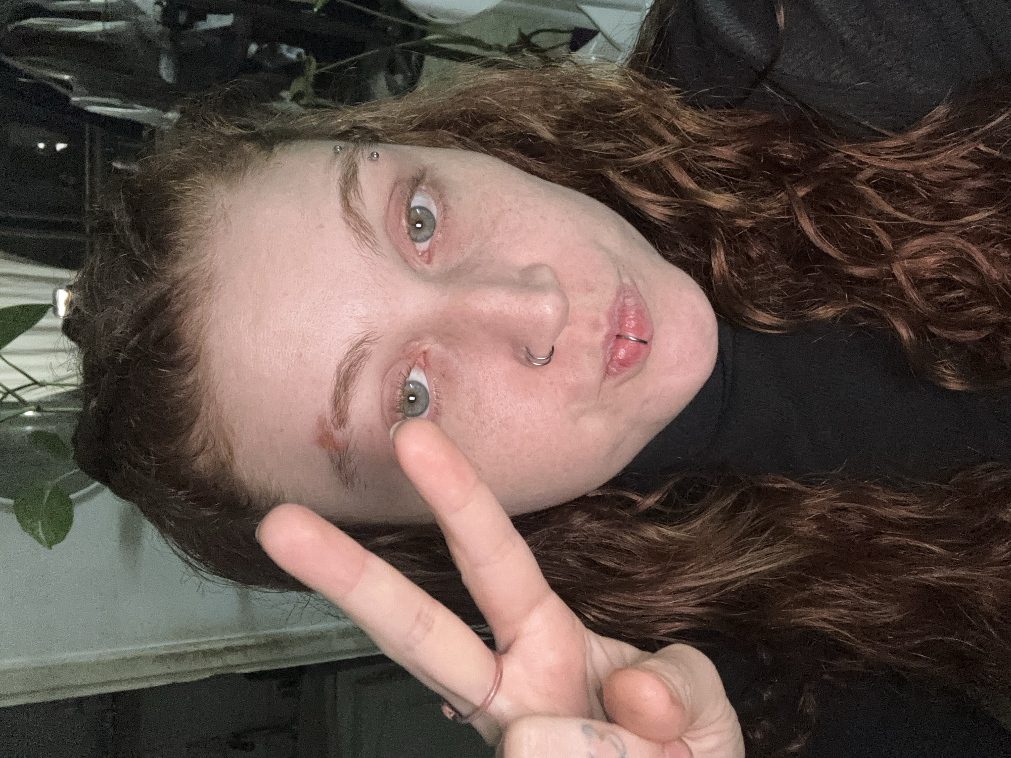 A person with auburn curly hair, fair skin, and visible facial piercings (eyebrow, nose, lip) is making a peace sign. The person is wearing a black turtleneck. The background appears to include a window and some plants. The location in the city of Buenos Aires, Argentina cannot be determined from the image.