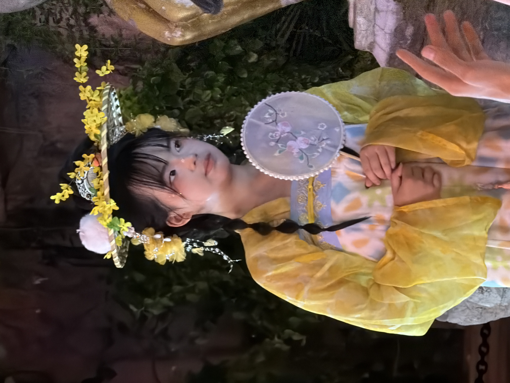 A young East Asian woman with dark hair styled in braids and adorned with yellow flowers and a fluffy white pom-pom, gazes to her right while holding a circular fan with a floral design. She is wearing a yellow hanfu-style garment with flowing sleeves over a patterned dress. Her face has light makeup, including pink blush on her cheeks. The background is dark and appears to be an outdoor setting with foliage and stone elements, possibly in Yuzhong District, China.