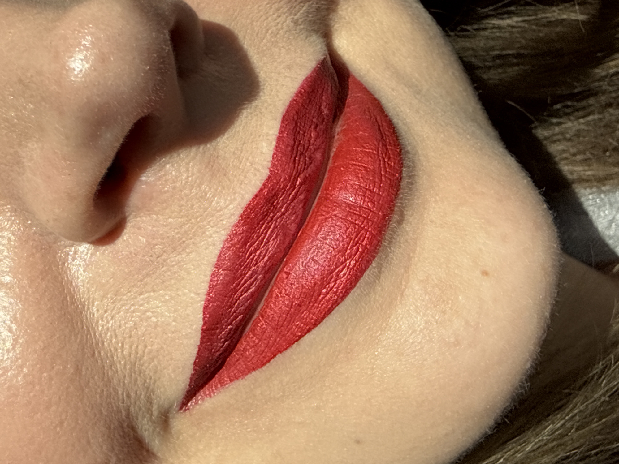 This close-up image, taken in Izhevsk, Russia, focuses on a person's mouth and nose area, highlighting vibrant red lipstick. The subject's lips are fully covered in a rich, matte or semi-matte red lipstick, displaying the natural texture of the lips with visible fine lines. The skin around the mouth and nose appears to be naturally shiny or slightly oily, with visible pores and fine vellus hair (peach fuzz). A small brown mole or freckle is discernible on the cheek to the right.
The scene is illuminated by bright, directional natural light, likely sunlight during the daytime, which casts clear shadows and creates prominent highlights on the skin's surface, particularly on the nose and upper lip area. Dark brown, possibly wavy, hair is visible along the right edge of the frame, slightly out of focus, suggesting it belongs to the person whose face is being shown.
The image does not depict any specific activity beyond showcasing the applied lipstick, possibly for a beauty or cosmetic purpose. There are no overt emotional expressions visible due to the extreme close-up. No text is visible in the image.