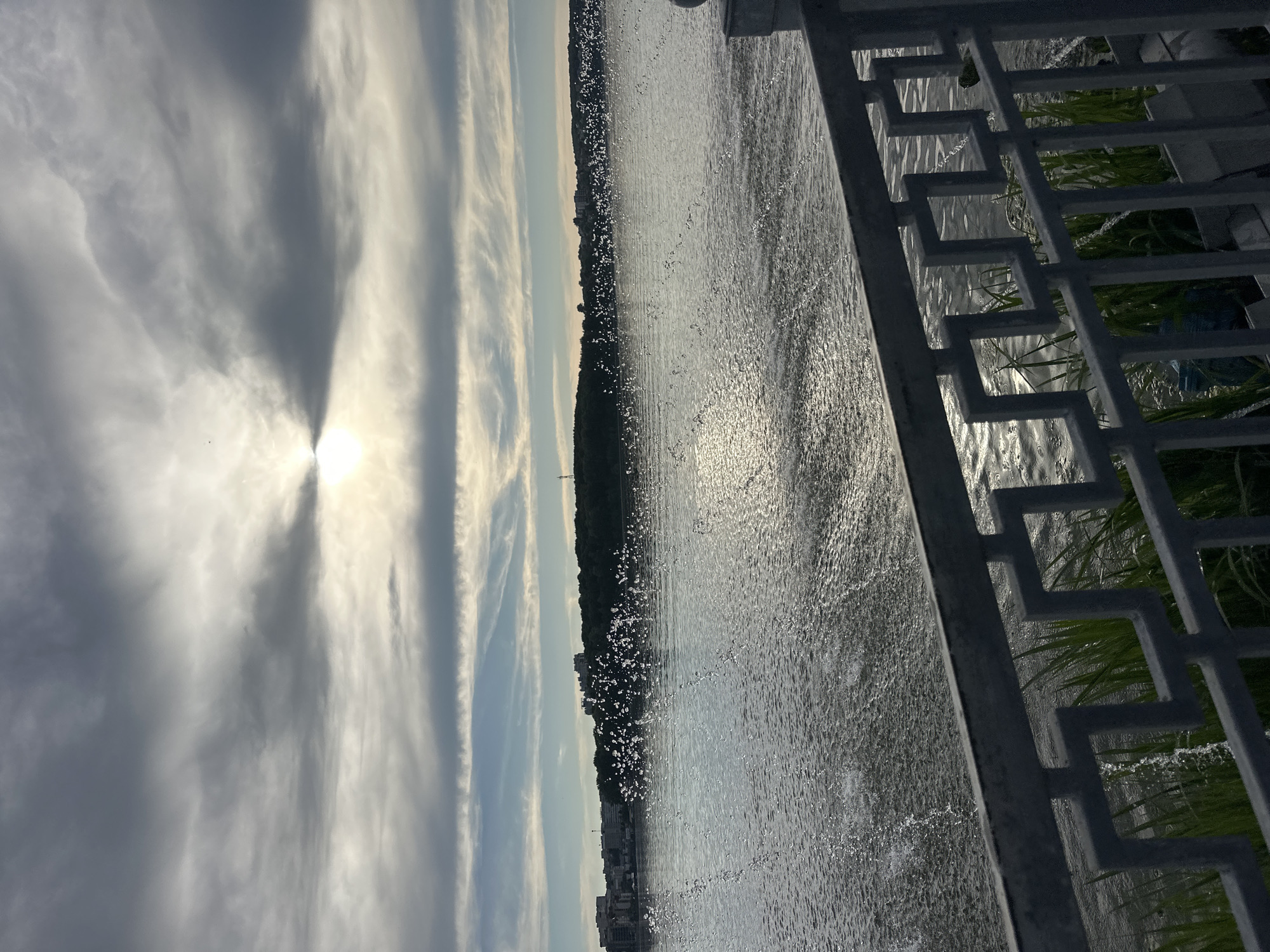 This image depicts a waterfront scene with a fountain in the foreground, located along the Ternopil pond in Ternopil, Ukraine. The sun is partially obscured by clouds, creating a diffused light that reflects off the rippling water. A metal railing with a geometric pattern runs across the bottom of the frame, separating the viewer from the pond. Beyond the fountain's spray, a tree-lined shore is visible, with buildings dotting the horizon. The sky is a mix of grey and blue clouds, with streaks of white suggesting movement. The overall atmosphere is calm and serene, with the gentle motion of the water and the distant cityscape creating a picturesque view.