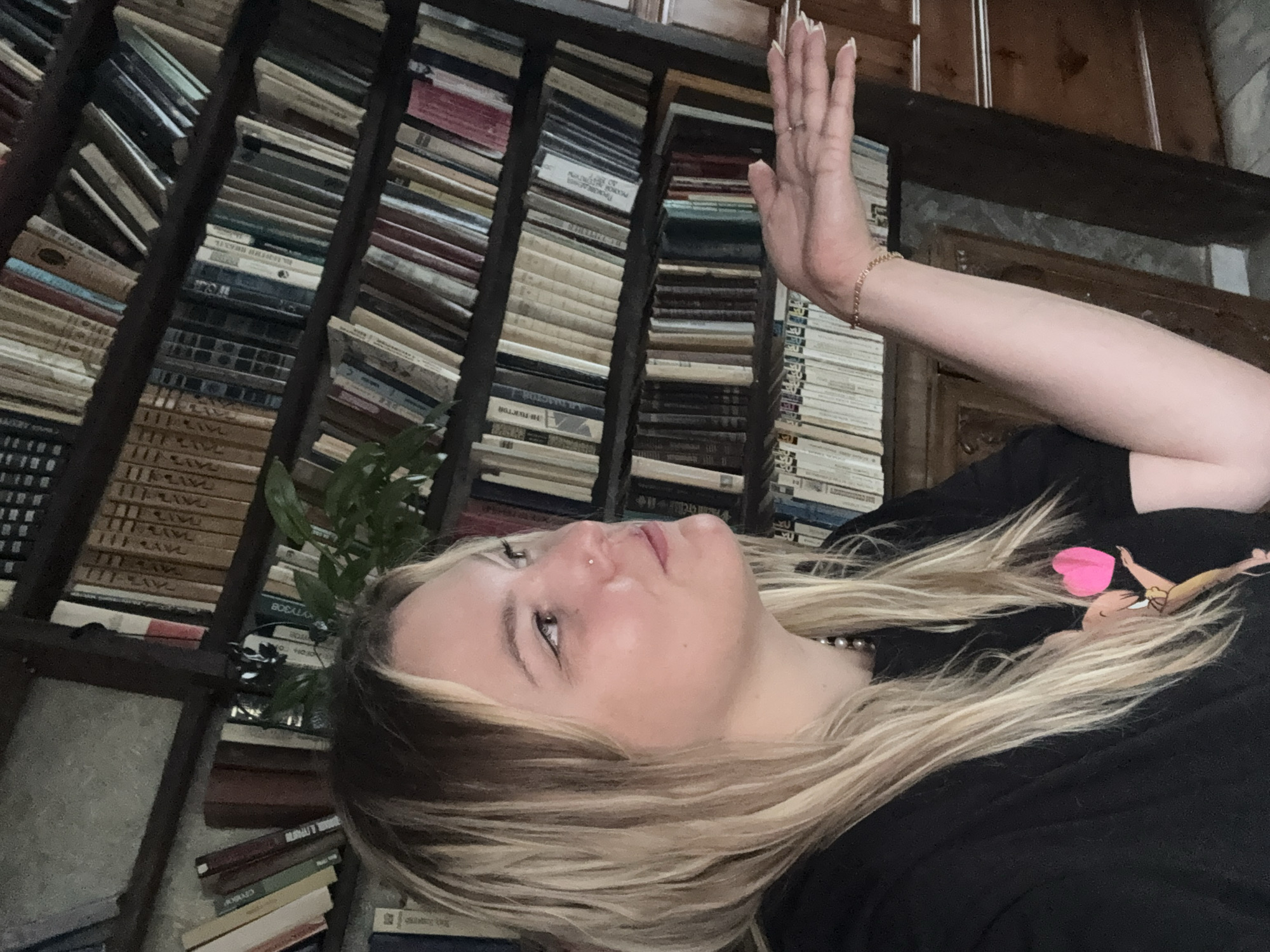 A young Caucasian woman with blonde hair and fair skin is captured in a medium shot, angled to her right, with a bookshelf filled with numerous books forming the background. She is wearing a black t-shirt with a graphic print of a cartoon character and a pink heart. Her right arm is extended, palm open, as if presenting something, and she is wearing a delicate gold bracelet on her wrist, along with a ring on her ring finger. Her facial expression is neutral, with her gaze directed off-camera. The environment suggests an indoor setting, possibly a library or a study, with the abundant collection of books indicating a scholarly or literary atmosphere. The lighting is moderate, with a slight warm tone. No specific location in Zavod Mikhailovskii, Russia, can be definitively identified from the visual information provided.
