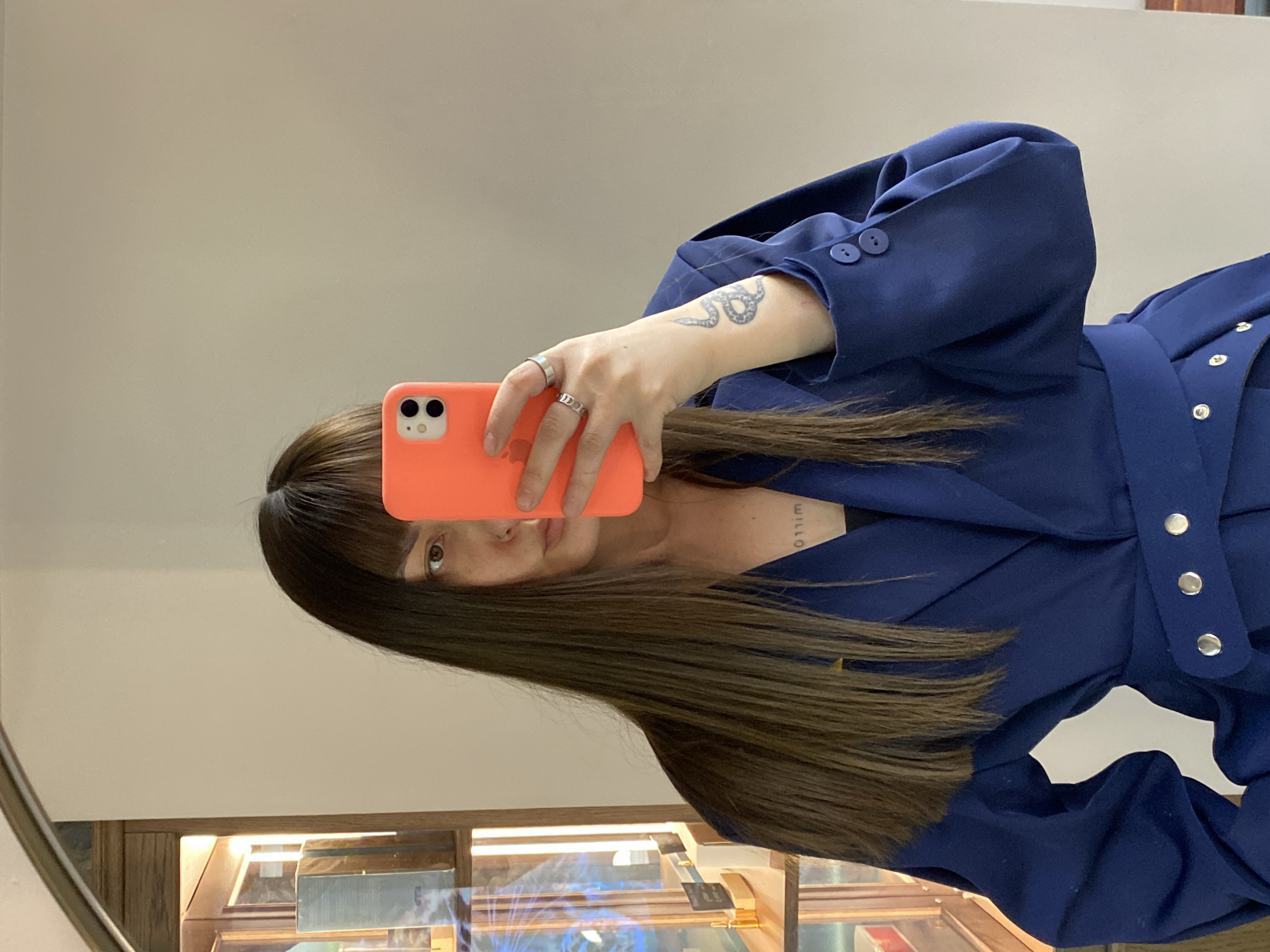 A young woman with long brown hair takes a selfie in a mirror. She holds an orange smartphone in her right hand, obscuring part of her face. She wears a navy blue blazer with decorative silver buttons and a self-tie belt. A tattoo is visible on her inner left arm. The reflection in the mirror shows the lower half of the woman's face, including her eye and lips. Behind her and reflected in the mirror is a display case containing various items, likely in a shop or store. The location is unspecified, but the city is identified as Votkinsk, Russia.