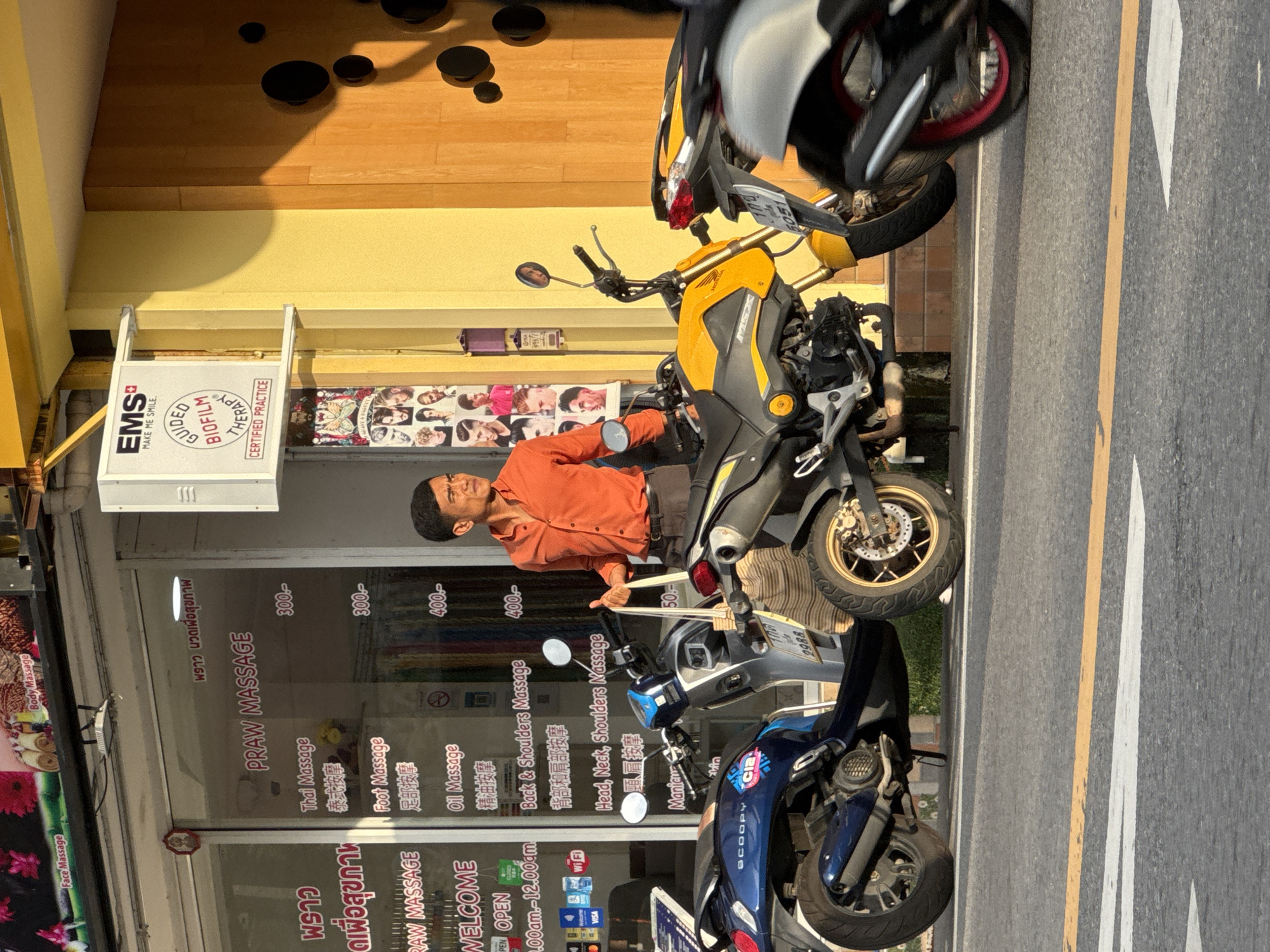 The image shows a street scene in Karon, Thailand, with a storefront for a massage business. The storefront has "PRAW MASSAGE" written in large letters on the glass. A price list for various massage services, such as "Thai Massage," "Foot Massage," "Oil Massage," and "Head, Neck, Shoulders Massage," is visible on the glass. A man in an orange shirt is standing next to the storefront, holding a bag. Several motorcycles are parked in front of the store, indicating a common mode of transportation in the area. The storefront appears to be a typical commercial establishment in a bustling Thai town. The sunlight suggests it is daytime. The overall environment is that of a street in a tropical city.