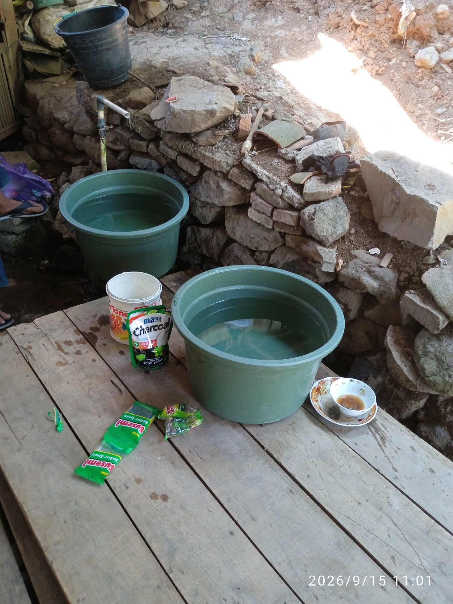 In a rustic outdoor setting, two large green tubs filled with water sit on a wooden platform. To the left, a black bucket is perched on a stone wall, with a water pipe and faucet above it. A white bucket and a pouch of "Mama Charcoal" dishwashing liquid are placed between the tubs. Several green packets of "Susemi" and a small dish with a brown liquid are also visible on the platform. The background consists of a stone wall and a dirt ground with scattered rocks, suggesting a simple, utilitarian setup, possibly for washing or general cleaning.