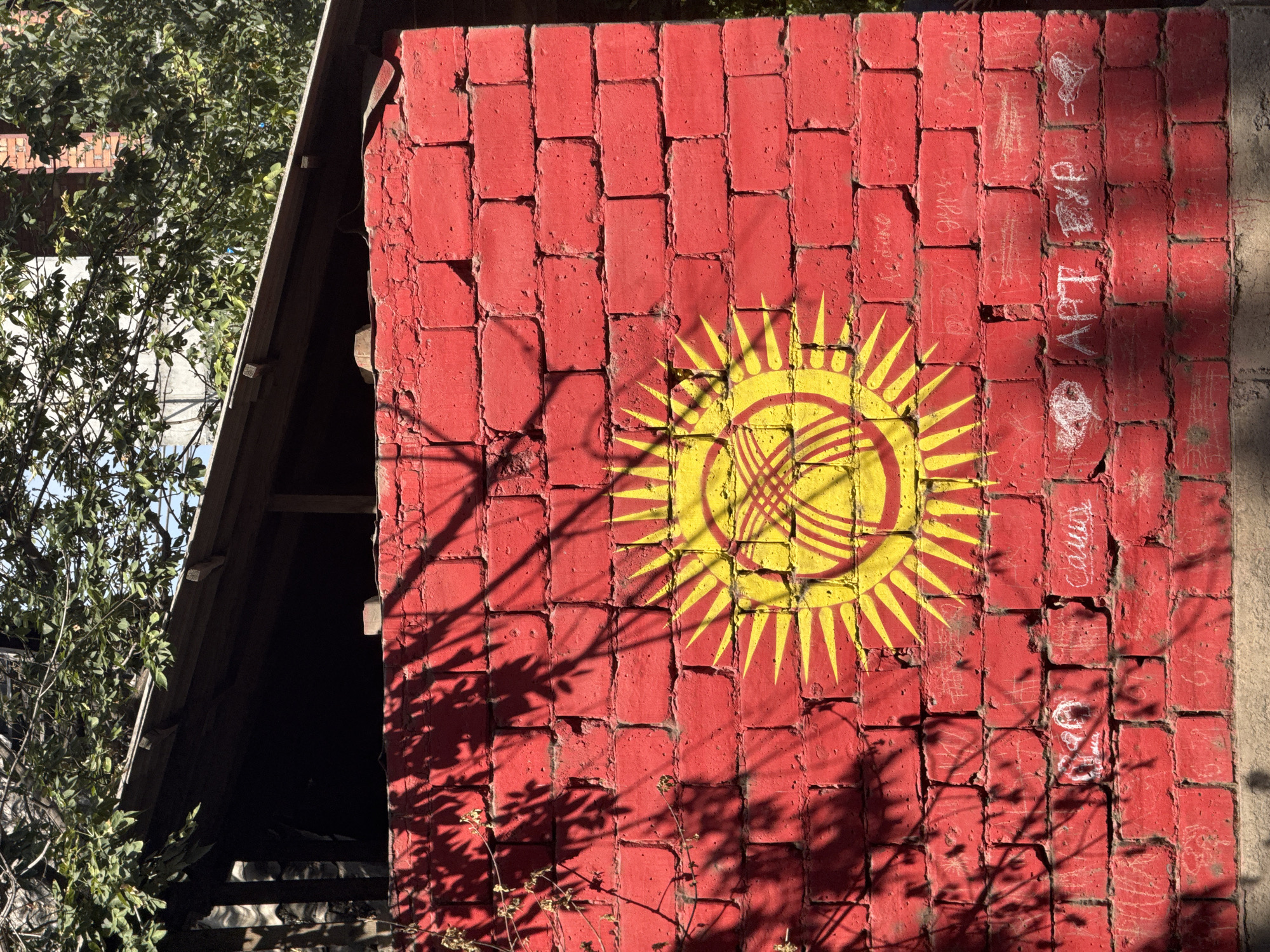 A close-up, eye-level shot captures the flag of Kyrgyzstan painted on a red brick wall. The flag features a stylized yellow sun with a central emblem. Shadows of tree leaves are cast across the surface of the wall, suggesting an outdoor setting. The brickwork shows signs of age and some graffiti is present on the lower portion of the wall. A wooden overhang, partially obscured by greenery, is visible at the top of the image.