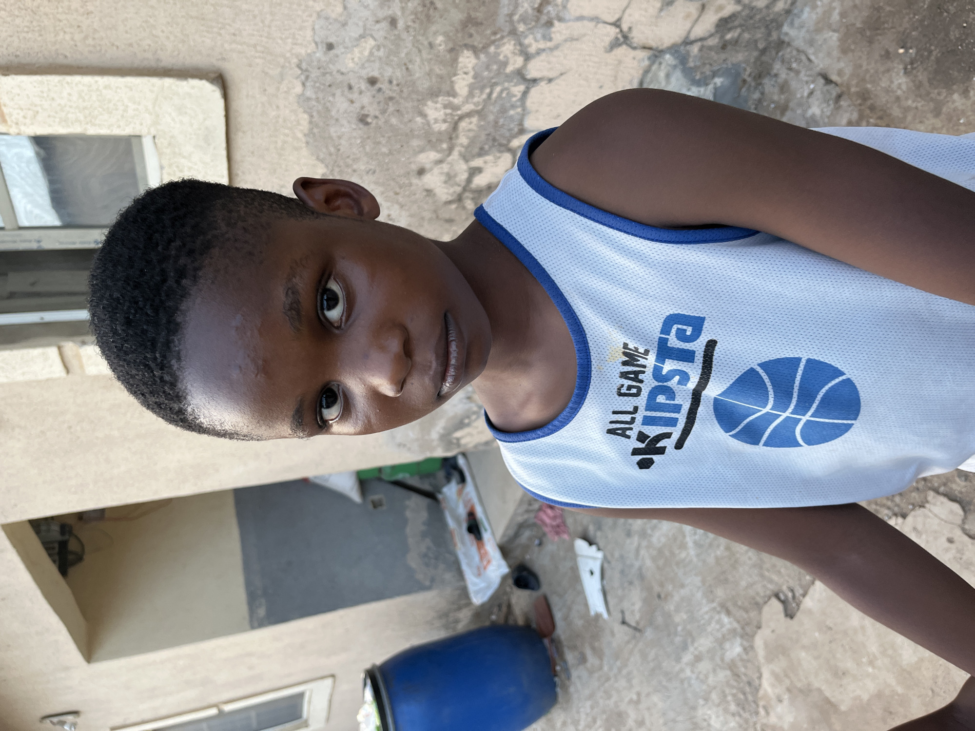 A young person stands in front of a building in Madalla, Nigeria. The individual has short, dark hair and brown skin, and is wearing a white basketball jersey with blue trim, a blue basketball design, and the word "KIPSTA" printed on it. The building behind the person is beige with peeling paint and has a small window. A blue barrel, white debris, and a white bag are at the edge of the building. The ground appears to be made of concrete.