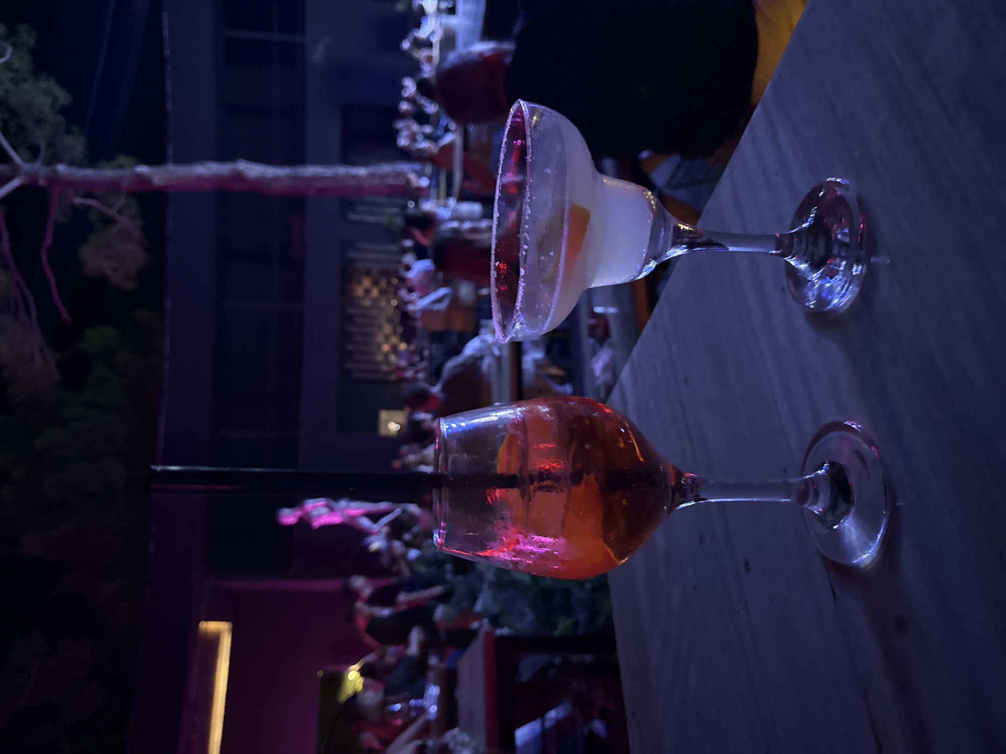 The image depicts a nighttime scene at an outdoor establishment, likely a bar or restaurant, in Bodrum, Turkey. Two cocktails are prominently featured in the foreground, placed on a wooden table. The cocktail on the left is in a wine glass, with a red-orange liquid, ice, a slice of orange, and a black straw. The cocktail on the right is in a margarita glass, with a pale liquid, a slice of orange garnish, and a salt-rimmed glass.
In the background, a crowd of people are visible, seated at tables and socializing. The lighting is dim, with purple and pink hues cast by ambient lights, suggesting a vibrant atmosphere. A tall tree trunk is visible in the middle ground, illuminated by the lights. Beyond the crowd, the architecture of the establishment, with dark structures and illuminated windows, is discernible. The overall impression is one of a lively evening gathering at a waterfront or popular nightlife venue in Bodrum.