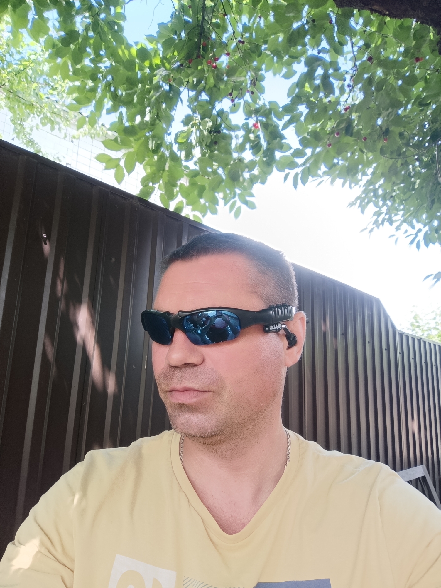 A man, wearing blue mirrored sunglasses and a light yellow t-shirt, is pictured from the chest up, with his head turned slightly to his left. He has short, dark hair and a light stubble on his face. The sunglasses have a small black attachment on the right side, resting near his ear. He is standing in front of a dark brown corrugated metal fence. Above and behind him, green leaves and a few red berries are visible, suggesting he is outdoors, possibly in a yard with a tree. The sky is bright and appears to be daytime. The setting is a suburban or residential area, consistent with the city of Ladyzhyn, Ukraine.