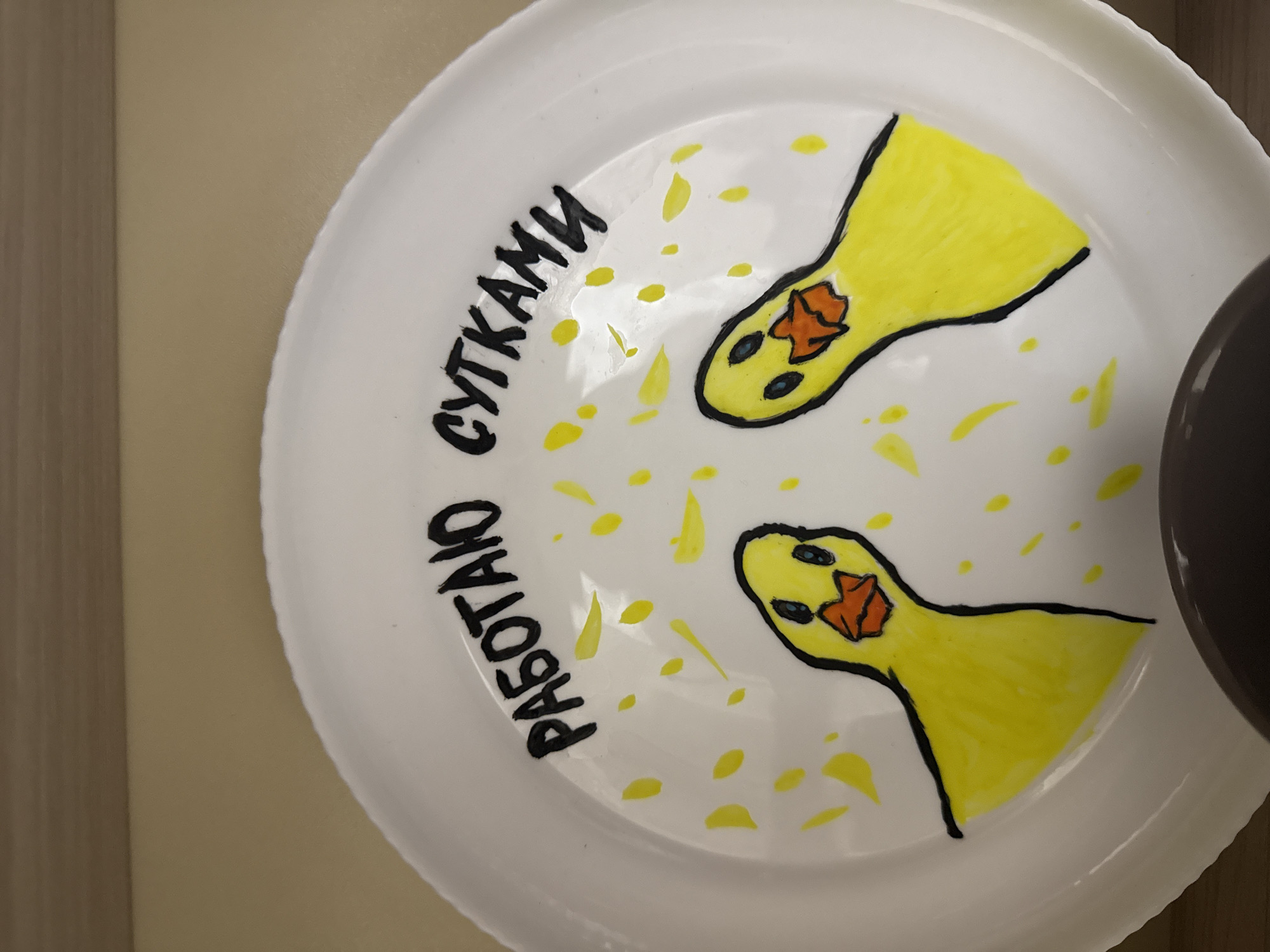 A white ceramic plate is depicted in the image, lying on a light brown surface. The plate features a hand-drawn design in black and yellow. Two cartoonish yellow ducklings with orange beaks and blue eyes are positioned on the plate. Scattered across the plate are small yellow dots and streaks, resembling confetti or paint splatters. Above the ducklings, in black lettering, are the words "РАБОТАЮ" (RABOTAYU) on the left and "СУТКАМИ" (SUTKAMI) on the right, which translate to "I work" and "around the clock" respectively. A dark circular object is partially visible at the bottom of the plate. The background consists of a plain light brown surface. The image is taken from a slightly elevated angle, looking down onto the plate.