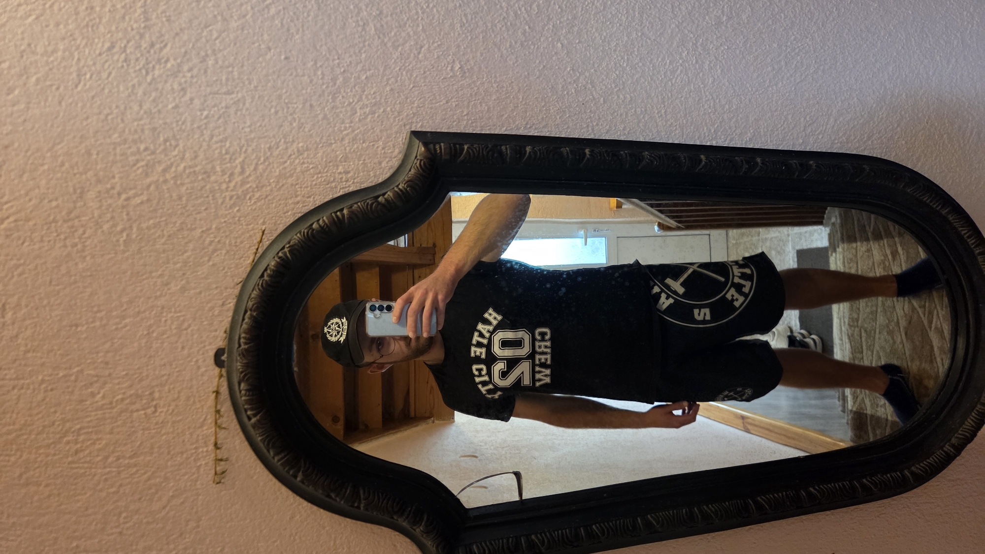 A man stands in front of a mirror in an indoor setting. He is wearing a black t-shirt with white lettering that reads "HATE CITY 05 CREW", a black baseball cap with a white emblem, black shorts with white circular logos, dark blue socks, and dark blue slip-on shoes. The man is holding a smartphone in his right hand, as if taking a selfie. The mirror is oval-shaped with a decorative black frame and is positioned against a light pink textured wall. In the reflection, a hallway with wooden stairs and a door is visible. The floor appears to be carpeted. There is some decorative twig-like object hanging on the wall above the mirror. The scene is lit with artificial light.