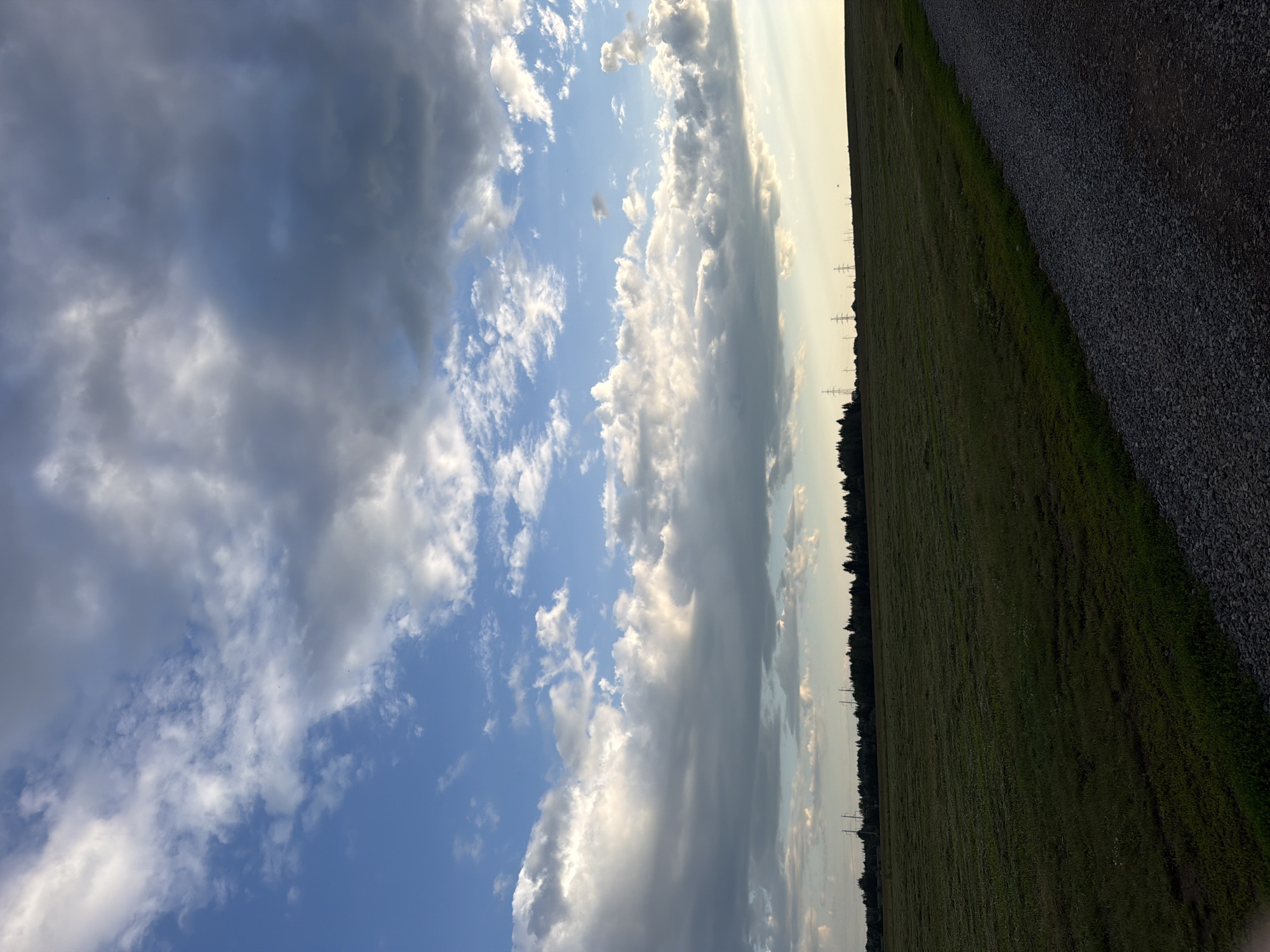 The image depicts a landscape scene under a partly cloudy sky, with the sun casting a warm glow. The horizon is visible, separating a vast expanse of clouds from a field of green grass. To the right of the field, a gravel path runs alongside it, suggesting a rural or outdoor setting. In the distance, a line of trees marks the edge of the field, with power lines and pylons visible interspersed along the tree line. The clouds are a mix of white and dark gray, indicating a dynamic weather pattern. The lighting suggests it is either early morning or late afternoon, given the soft, diffused sunlight. There are no people or animals visible in the frame. The overall impression is one of natural tranquility and open space.