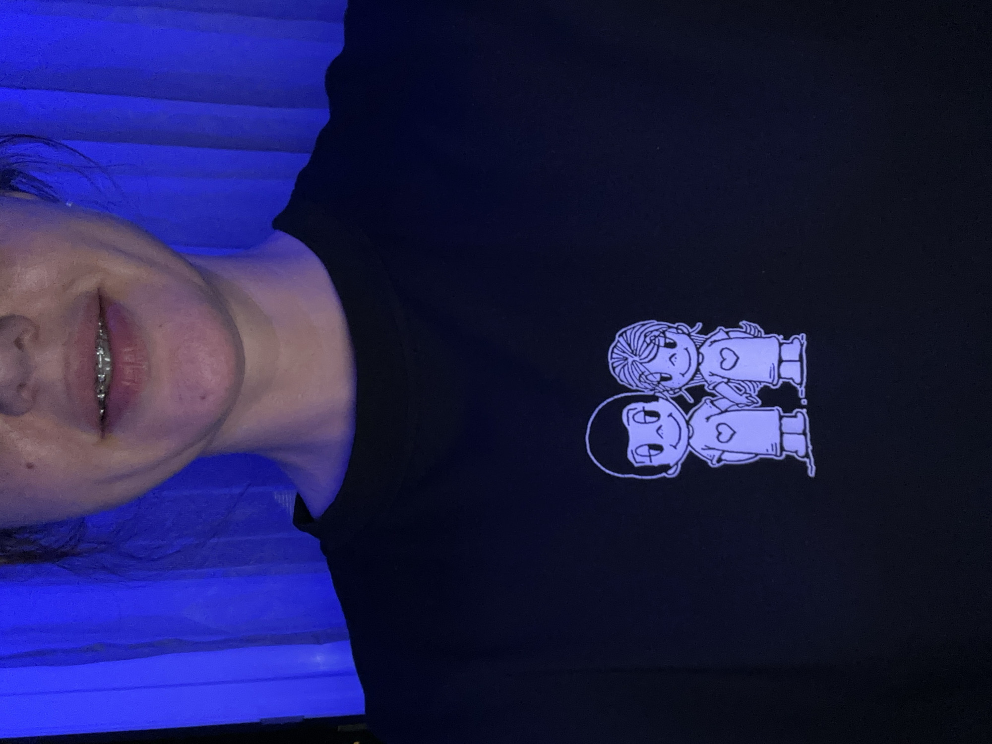 The image shows a close-up of a person wearing a black t-shirt with a white cartoon illustration of two figures holding hands. The figures are stylized, with simple features and clothing, and each has a heart shape on their chest. The person's mouth is visible, with braces on their teeth. The background is a dark blue, possibly curtains, illuminated by a blue light. There is no indication of the city of Sochi, Russia, in this image.