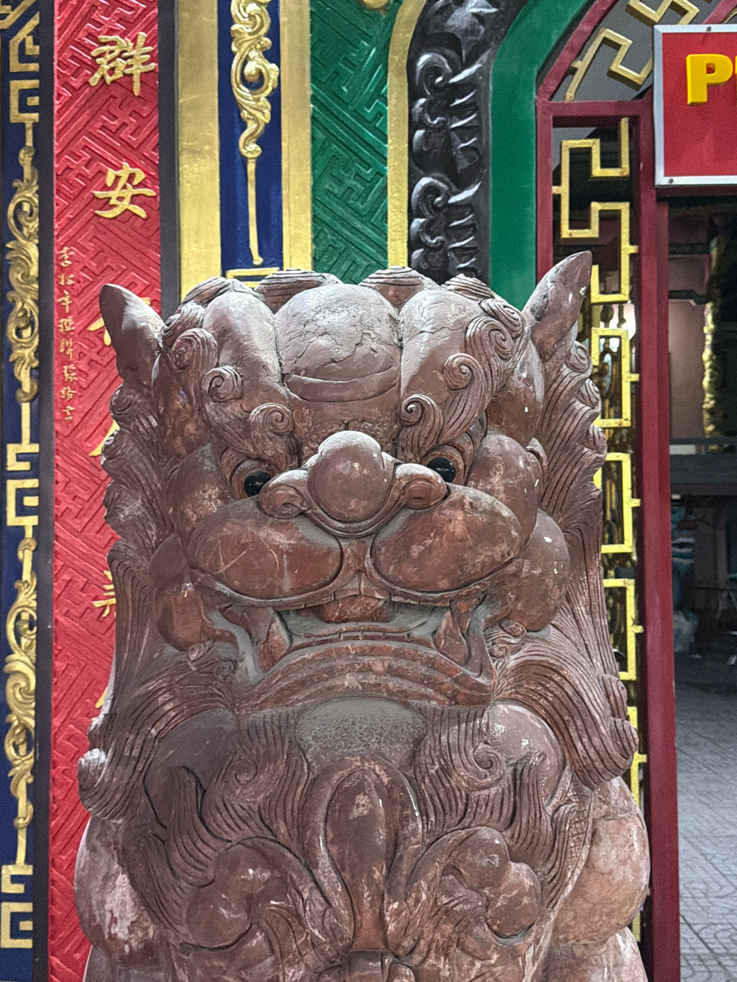 A close-up, eye-level, medium shot captures a weathered, reddish-brown stone statue of a mythical beast, possibly a guardian lion, positioned outdoors in front of a vibrantly decorated wall. The statue is the primary subject, its intricately carved features, including a scaly mane, prominent eyes, and a gaping mouth, dominating the foreground. The beast's head is tilted slightly forward, with its gaze directed towards the viewer.
Behind the statue, a colorful, vertical pattern of decorative panels is visible. The left panel features a red background with intricate gold Chinese characters that appear to be "群安" (Qún Ān), meaning "Peaceful Group" or "Peaceful Crowd," and smaller characters below. Next to this is a blue panel with a vertical gold ornamental motif. To the right of the blue panel is a textured green panel with a repeating geometric pattern. Further to the right, a red pillar frames a section of a building with a gold, lattice-like design and a red sign with a yellow letter "P" on it. The ground beneath is paved with light-colored square tiles. The lighting suggests an outdoor setting during the day, with some shadows indicating it's not direct overhead sun. The overall impression is one of cultural ornamentation and a protective, mythical presence.
This statue is likely located in Phú Quốc, Vietnam, a popular tourist destination known for its temples, pagodas, and ornate architecture, which often feature such guardian statues.