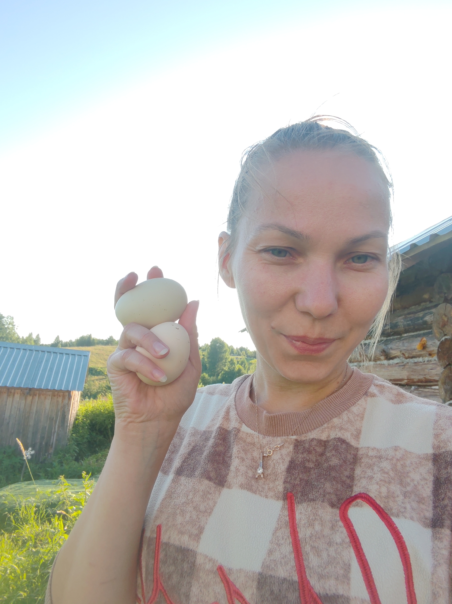 A blonde woman with blue eyes is taking a selfie outdoors in a rural setting, holding two light-colored eggs in her left hand. She is wearing a light-colored, fuzzy, plaid sweater with red embroidery. A thin necklace with a small Eiffel Tower pendant is visible. The background shows a wooden building, a shed with a metal roof, green vegetation, and a clear sky. The image appears to have been taken on a sunny day, possibly in the city of Chuchlem, Russia, given the architectural style of the buildings and the natural landscape.