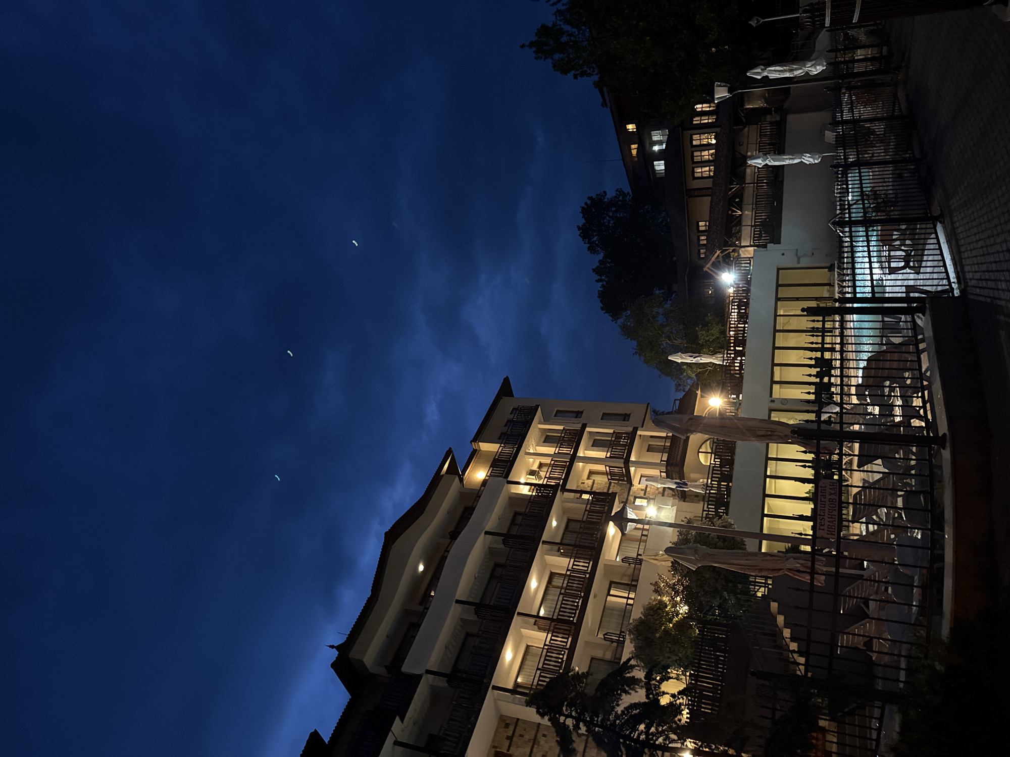 The image shows a nighttime view of a hotel or resort in Varna, Bulgaria. The sky is a deep blue with faint clouds and a crescent moon. Two multi-story buildings are visible. The building on the left is illuminated with lights from the balconies. The building on the right has lighted windows and is partially obscured by trees. A black metal fence is positioned in the foreground, with glimpses of a swimming pool visible behind it. Umbrellas and outdoor furniture are also visible. A sign with "Restaurant Иванчов Хан" is seen on the fence.