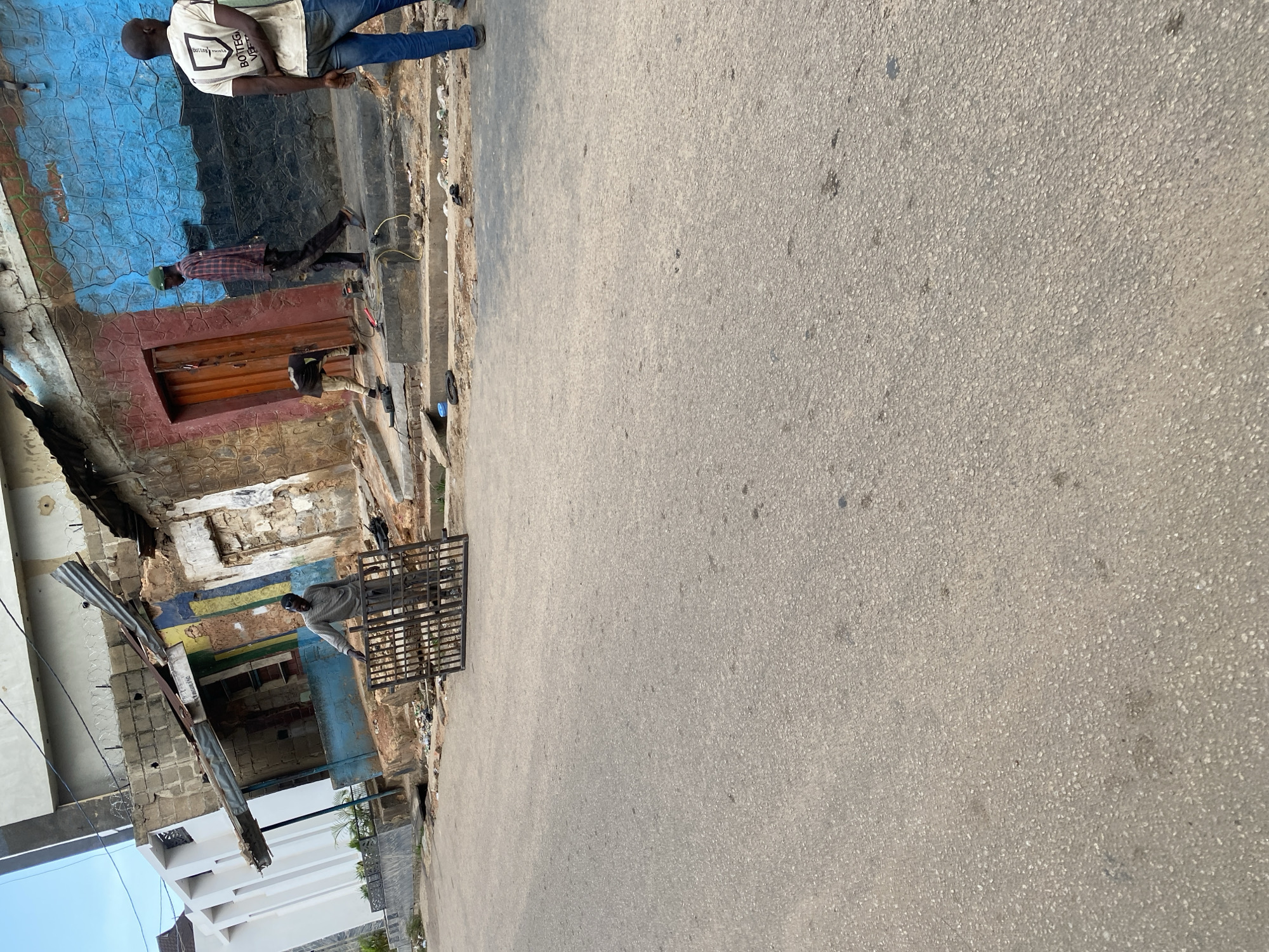 In Jos, Nigeria, this image captures a street scene with a focus on a dilapidated building. Three individuals are visible. One man, wearing a long-sleeved shirt and pants, stands near a metal gate on the street. Another man, in a plaid shirt and green cap, walks along the elevated sidewalk in front of the building. A third man, in a white t-shirt and blue jeans, stands facing away from the camera on the sidewalk.
The building exhibits signs of disrepair, with a corrugated metal roof extension and weathered walls. A colorful mural, featuring blue and red sections, adorns one part of the facade. Various tools and materials are present on the sidewalk, suggesting ongoing work or construction. The street itself is paved with asphalt, showing some wear and tear. The overall environment appears to be an urban setting, likely a commercial or residential area within the city.