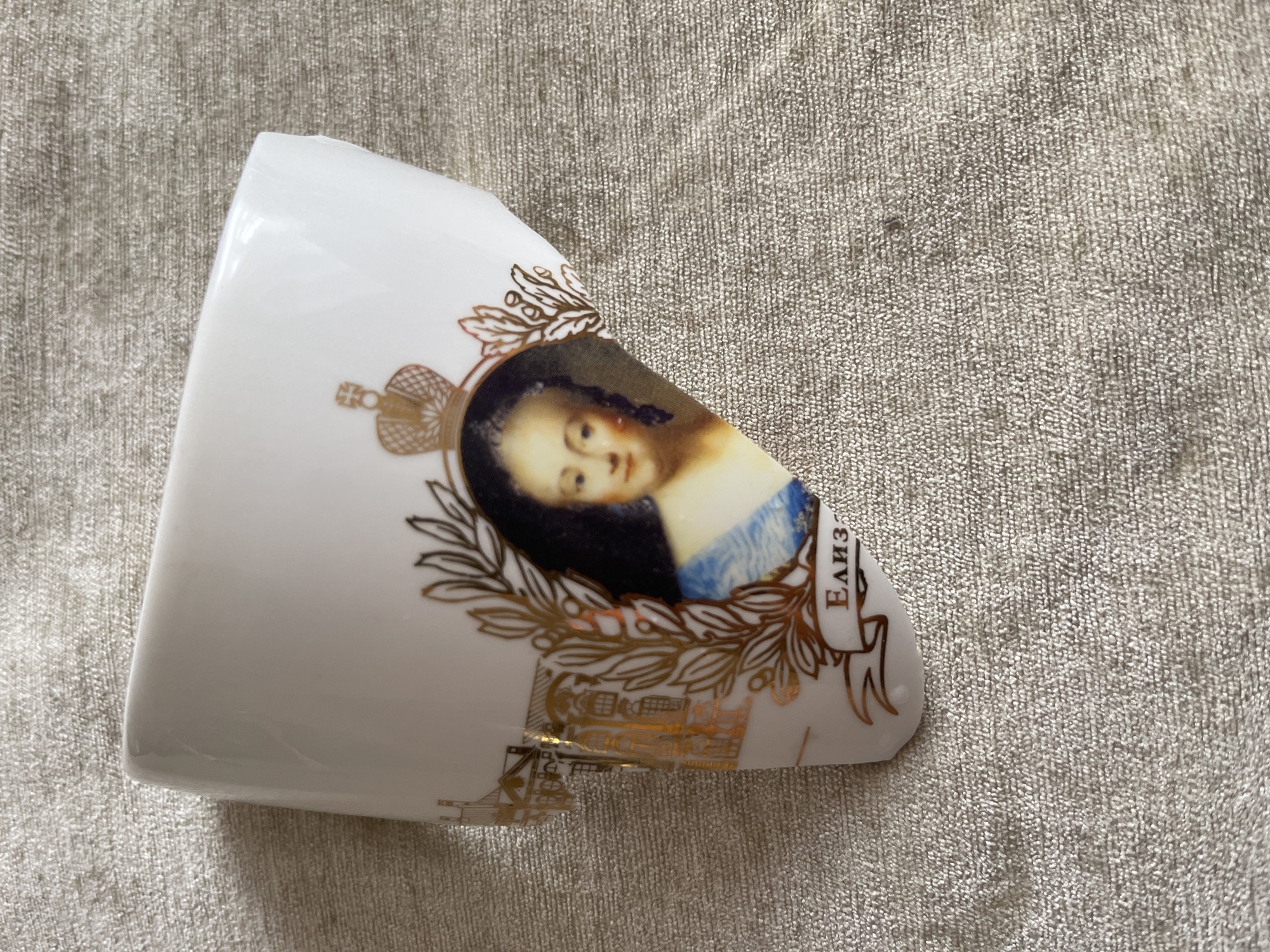 A fragment of a white porcelain cup rests on a textured, light-gray fabric. The cup fragment features a portrait of a woman with dark hair and fair skin, framed by golden laurel leaves and a crown. Adjacent to the portrait, script appears in a foreign language, reading "Елиз." Further gilded decorations adorn the cup fragment. The cup is broken and missing pieces. While the image includes a portrait of a crowned woman in a style reminiscent of Russian royalty, it does not provide enough information to pinpoint a specific location in Moscow.