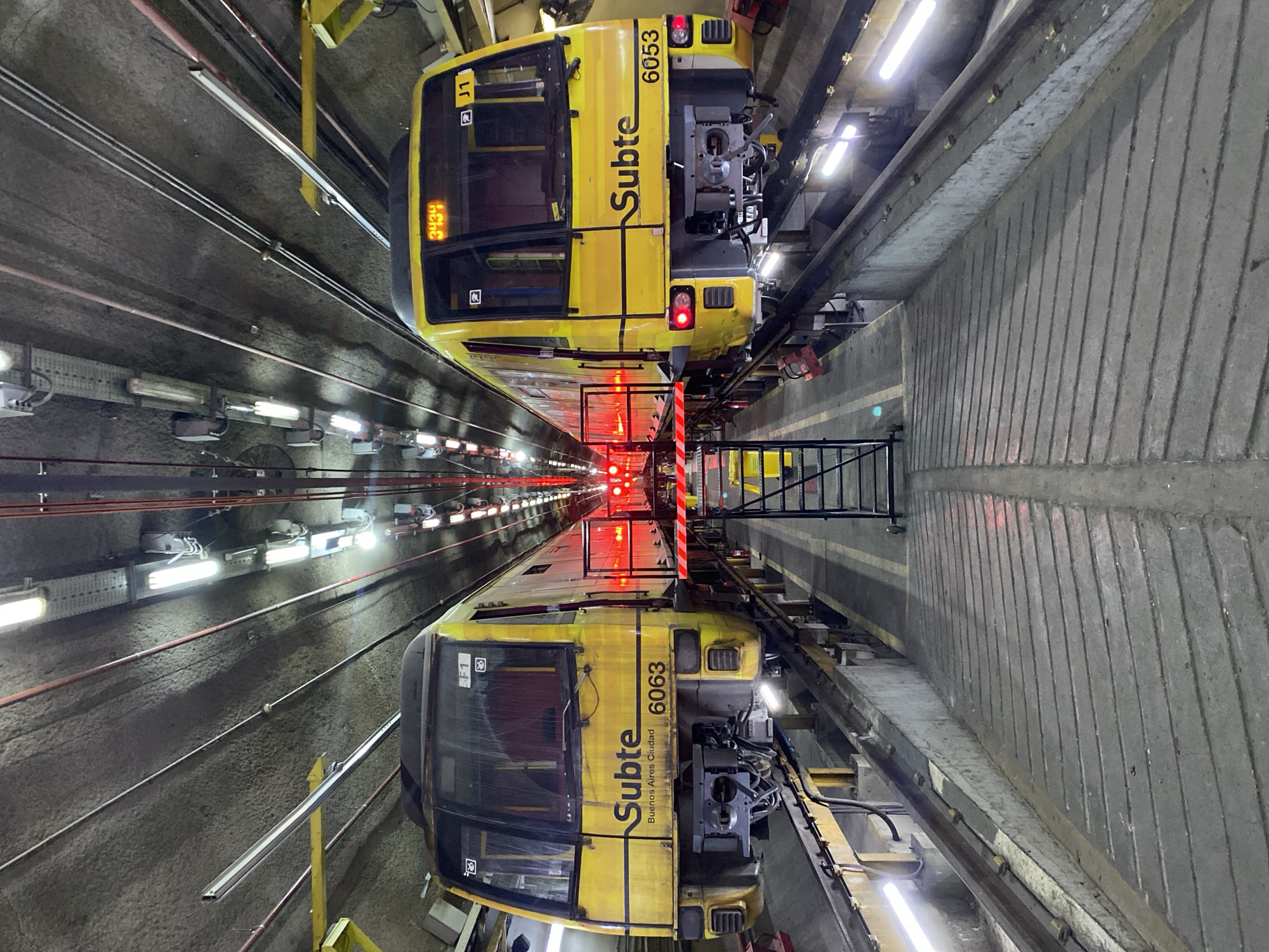The image depicts two yellow subway trains positioned in an underground tunnel or maintenance facility. The trains are parked on parallel tracks, facing each other, and are brightly illuminated by numerous fluorescent lights attached to the concrete walls and ceiling. Both trains prominently display "Subte" and "Buenos Aires Ciudad" on their sides, indicating their affiliation with the Buenos Aires subway system in Argentina.
The upper train is marked with the number "6053" and "J1" near its driver's cab, with a digital display showing "3434". The lower train is identified by the number "6063". Red lights glow from the front of both trains, and a series of additional red signal lights extends down the center of the tunnel. A dark metal ladder or platform stands between the trains near their front ends. The concrete tunnel walls feature a textured pattern of vertical grooves, and various electrical conduits, pipes, and cables run along them. Yellow and black striped safety barriers are also discernible.
No people are visible in the scene, suggesting the trains are stationary for maintenance, storage, or inspection. The artificial lighting confirms an enclosed, subterranean environment, offering a glimpse into the operational infrastructure of the Subte system.