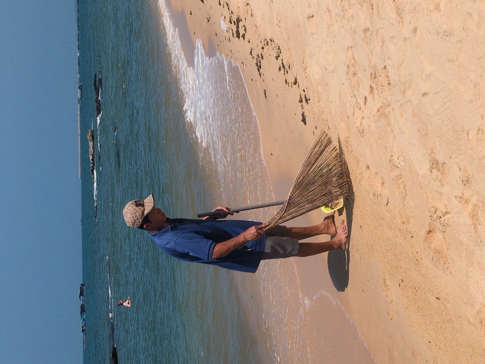 A man in a blue polo shirt and a tan patterned baseball cap sweeps sand on a beach near the ocean in Phú Quốc, Vietnam. He is barefoot and holding a long-handled broom. A small yellow object is visible near his feet, embedded in the sand. The beach is tan with wet areas near the water. The ocean is blue-green and contains some visible rocks and people. The sky is blue.