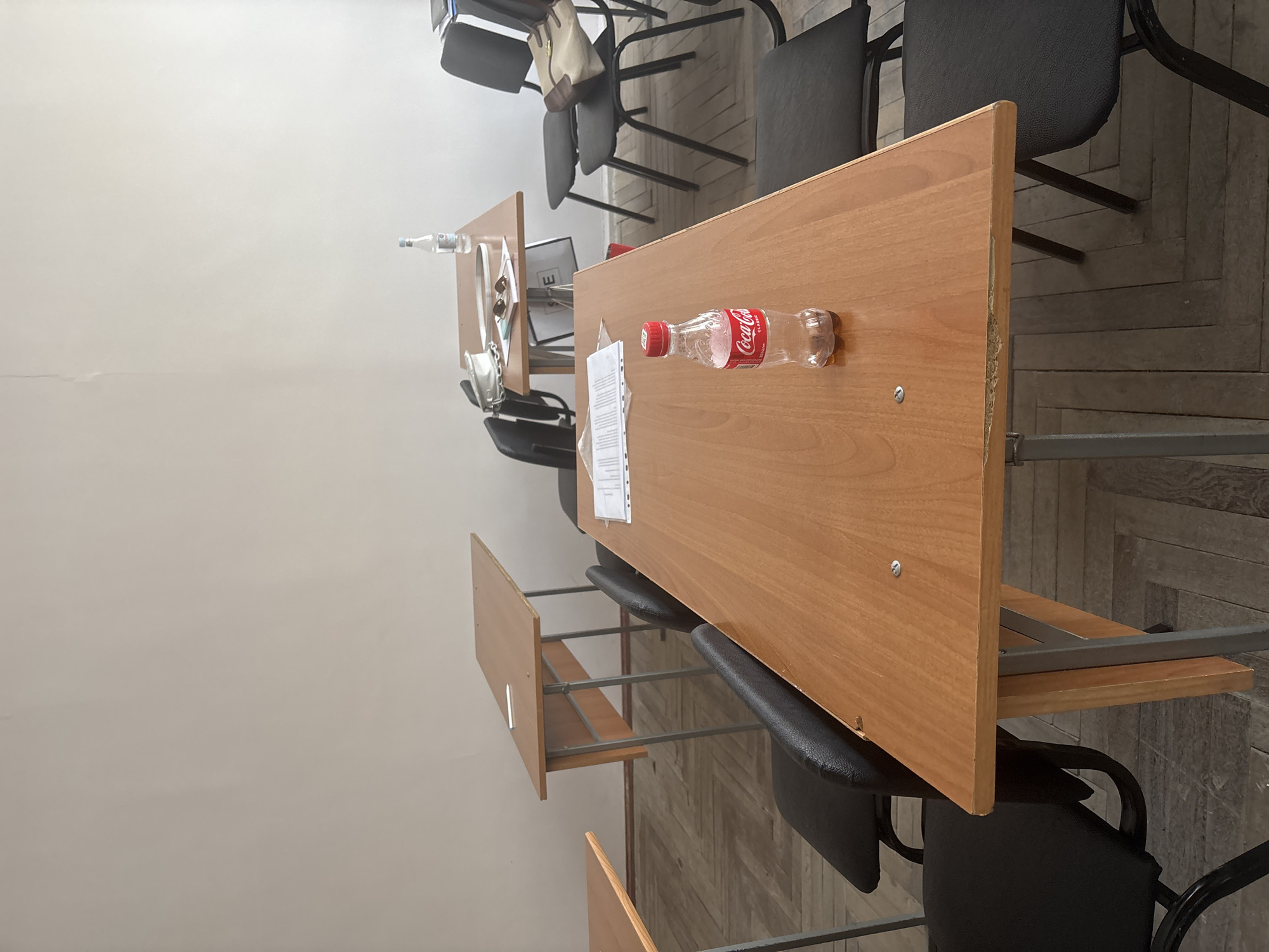 The image shows a classroom in Yerevan, Armenia. The room is furnished with rows of desks and chairs. On one of the desks, there is a bottle of Coca-Cola, a water bottle, and some papers. There are other desks visible, some with bags and other items on them. The walls of the room are plain, and the floor is tiled.