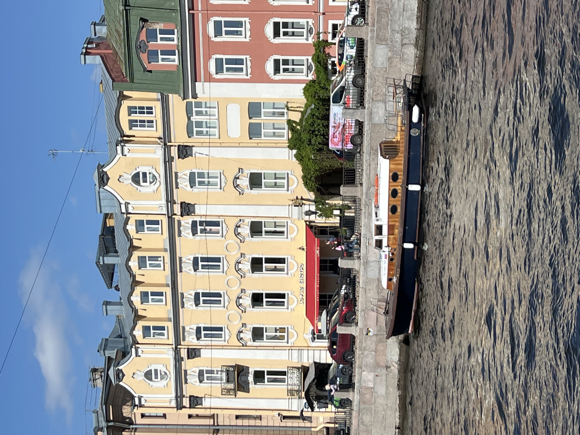 The image captures a riverside scene in Saint Petersburg, Russia, featuring a cluster of colorful buildings lining the Neva River. The buildings display a variety of architectural styles and colors, ranging from green and red to yellow and beige. Each building features numerous windows. A red awning extends from one of the buildings.
Along the riverbank, a concrete walkway runs alongside the river. A boat is docked on the river. Several vehicles, including vans and cars, are parked on the street adjacent to the buildings. A patch of greenery climbs the buildings. People are seen walking on the sidewalk in front of the buildings. The Neva River is textured and appears to be moderately turbulent. The sky is clear and blue.