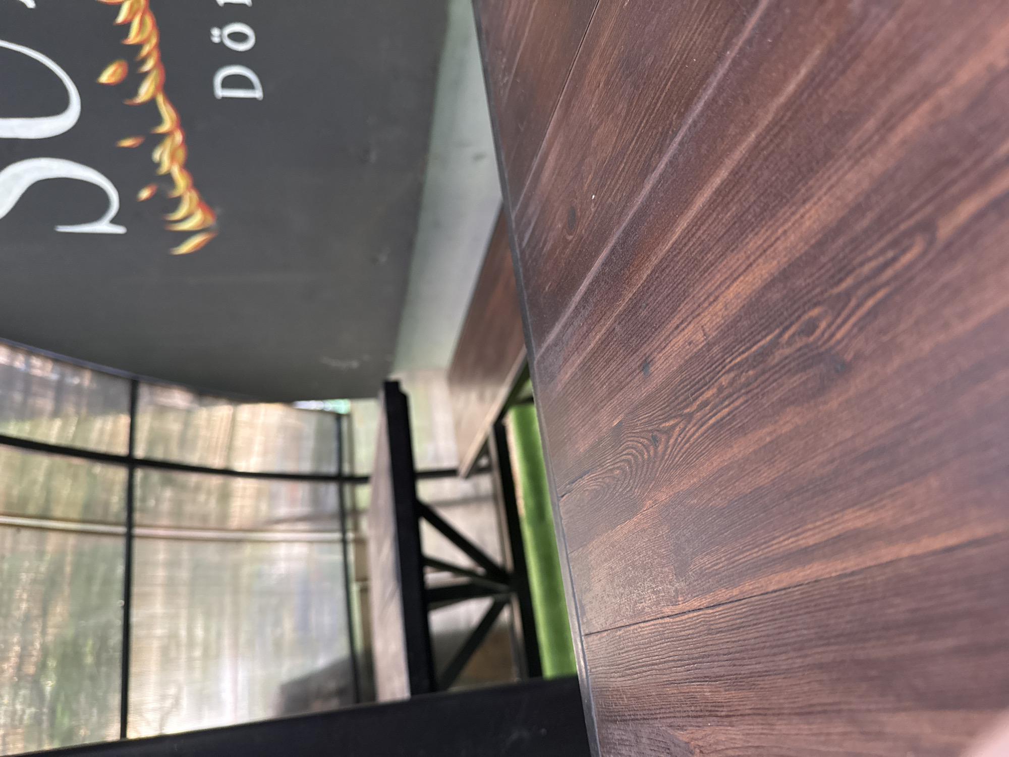 The image shows a close-up of a wooden table with a dark brown, wood grain finish. The table is set outdoors under a covered structure with a transparent, ribbed roof. In the background, another dark wooden table and bench are visible, along with green turf. To the right, a black wall features a white logo with flames and the word "Dön" in white letters. The overall scene suggests an outdoor seating area at a restaurant or cafe. There are no people visible in the image.